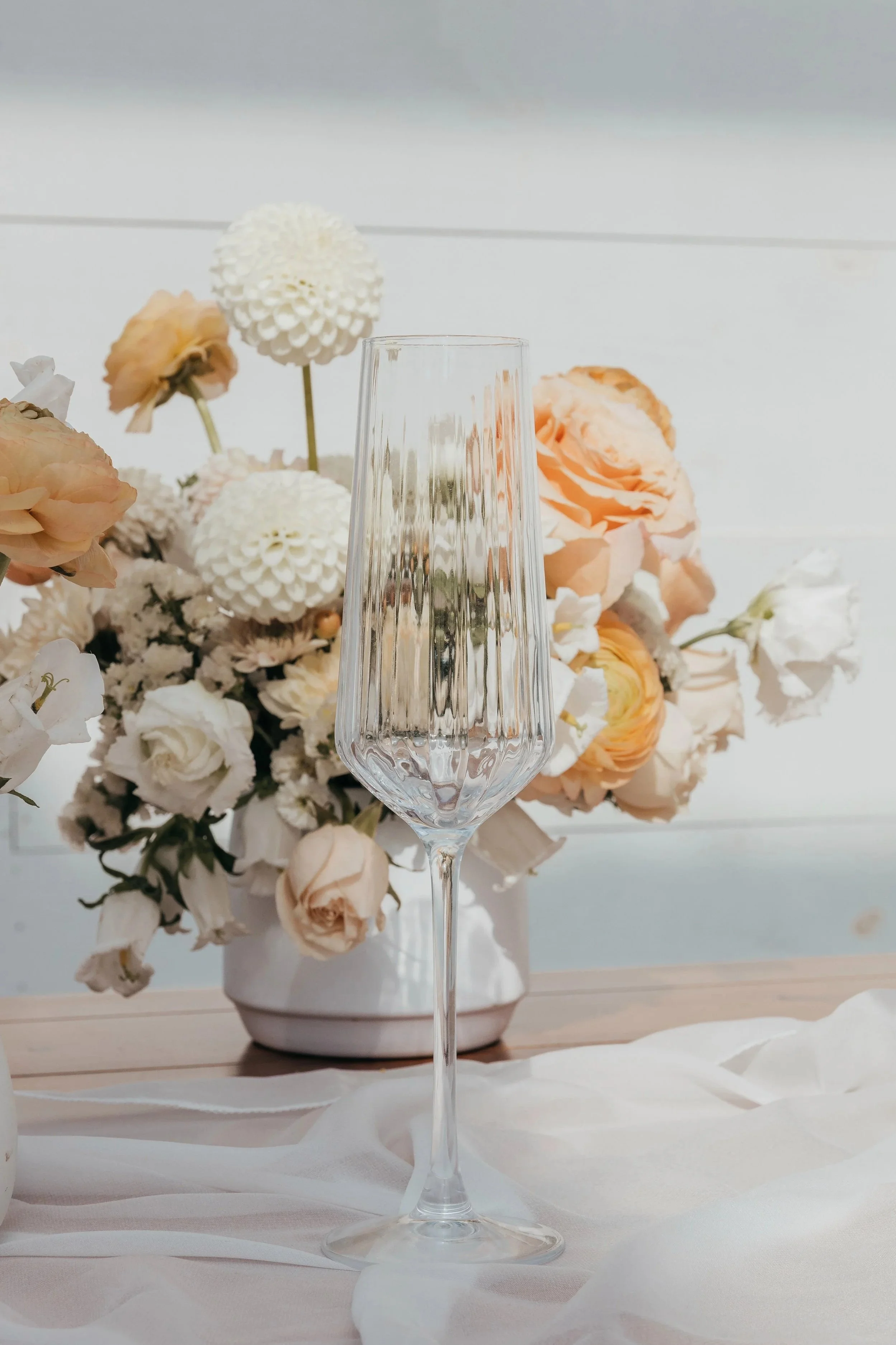 An empty crystal champagne flute on a table, with a bouquet of white and peach flowers in the background.