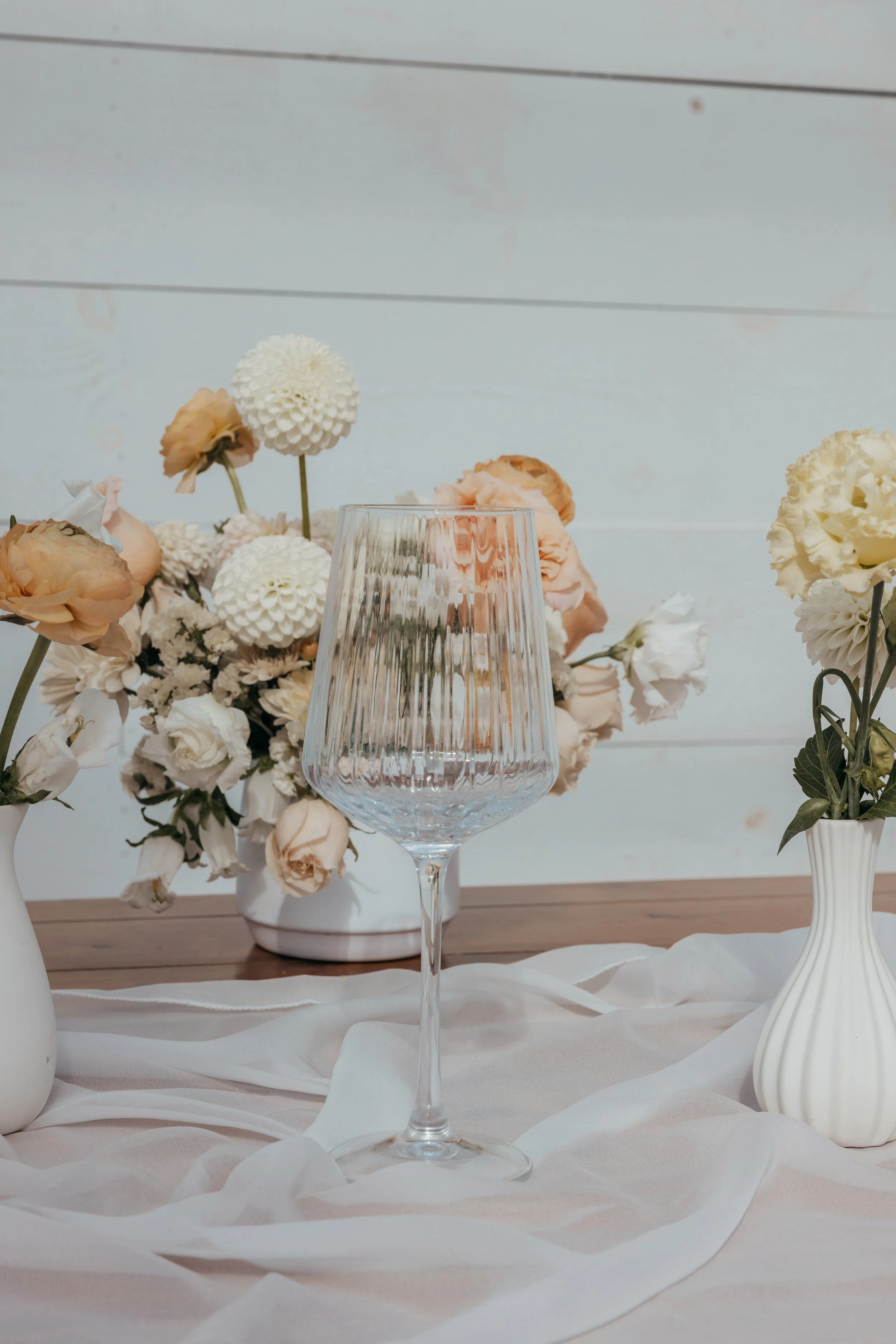 Empty wine glass on a table with white fabric, surrounded by vases with cream and peach flowers, and a light-colored wooden background.