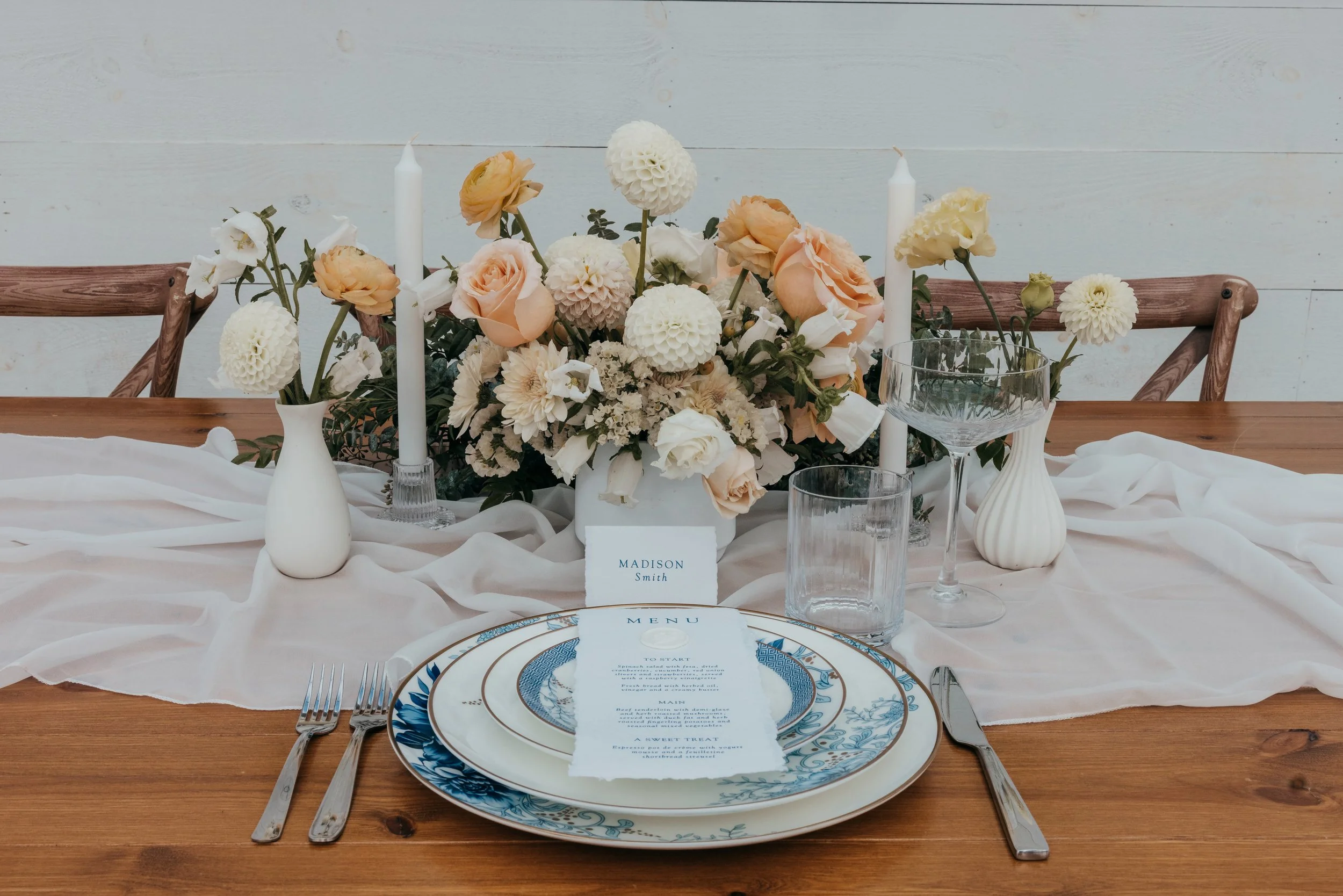 Elegant table setting with a floral centerpiece, white and blue patterned plates, silverware, and glassware, against a white backdrop with chairs visible.