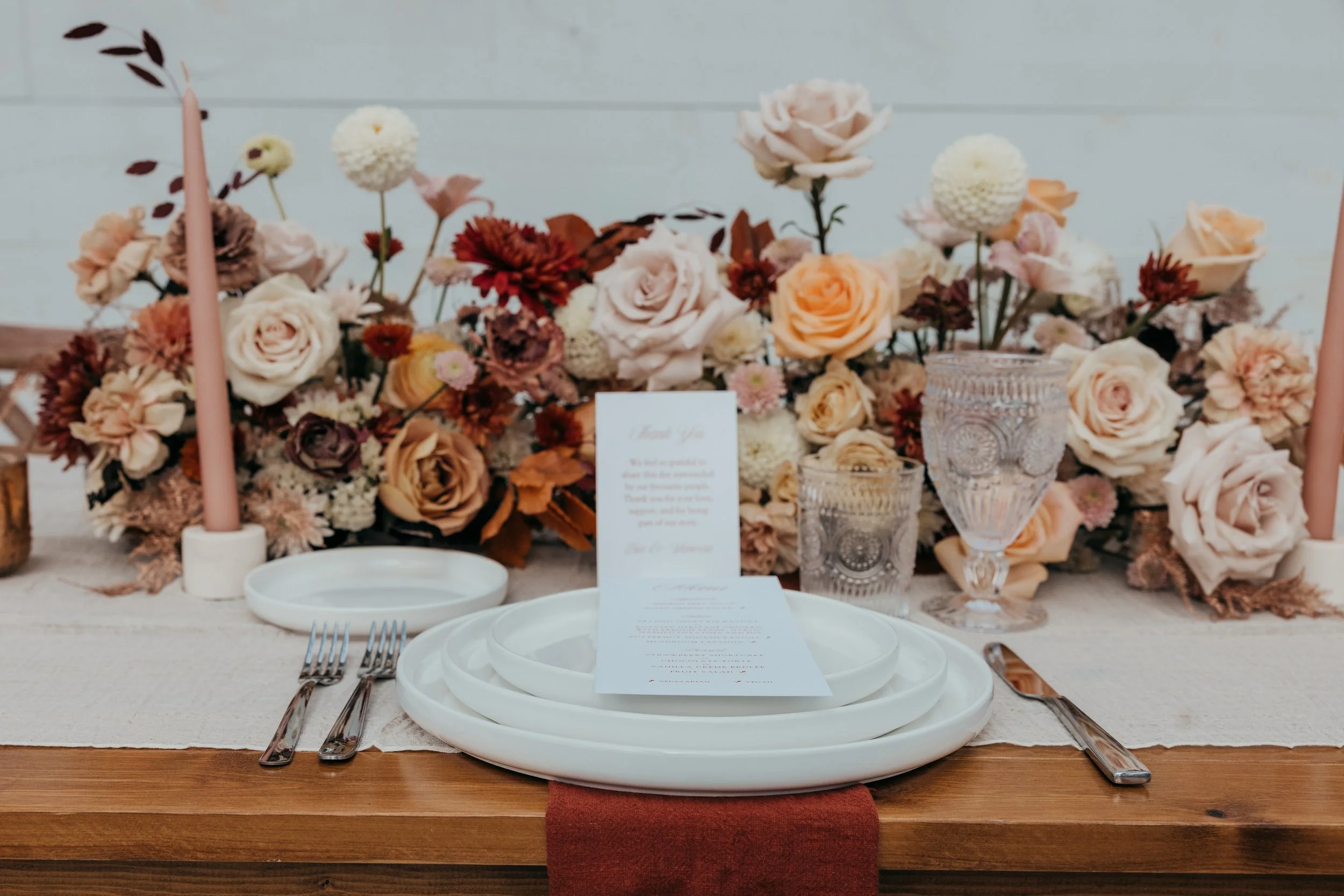 Elegant wedding table setting with white plates, silverware, pink candles, crystal glasses, and a floral centerpiece with roses and other flowers in soft colors.