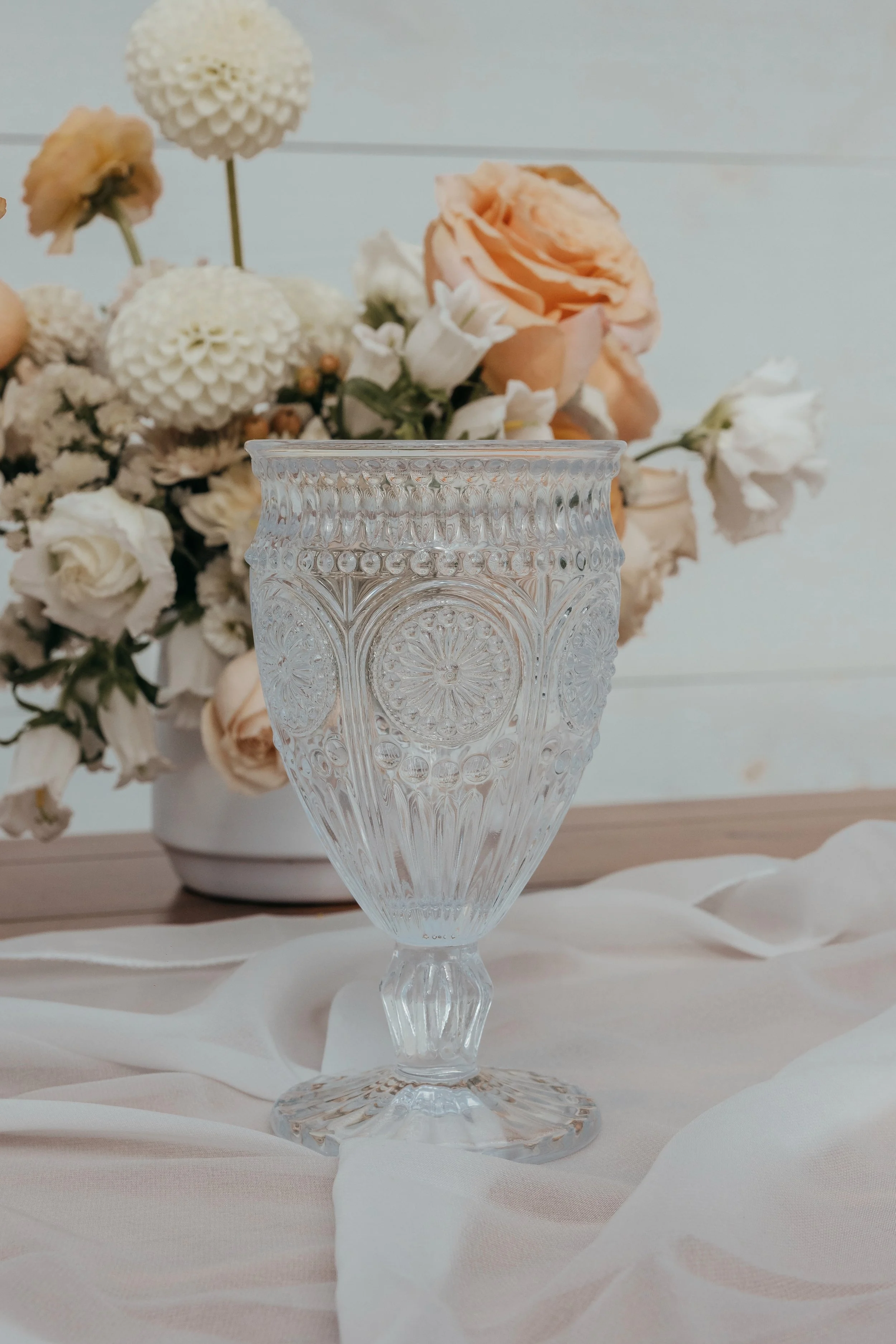 Elegant cut glass vase on a white fabric with a floral arrangement of blush and white flowers in the background.