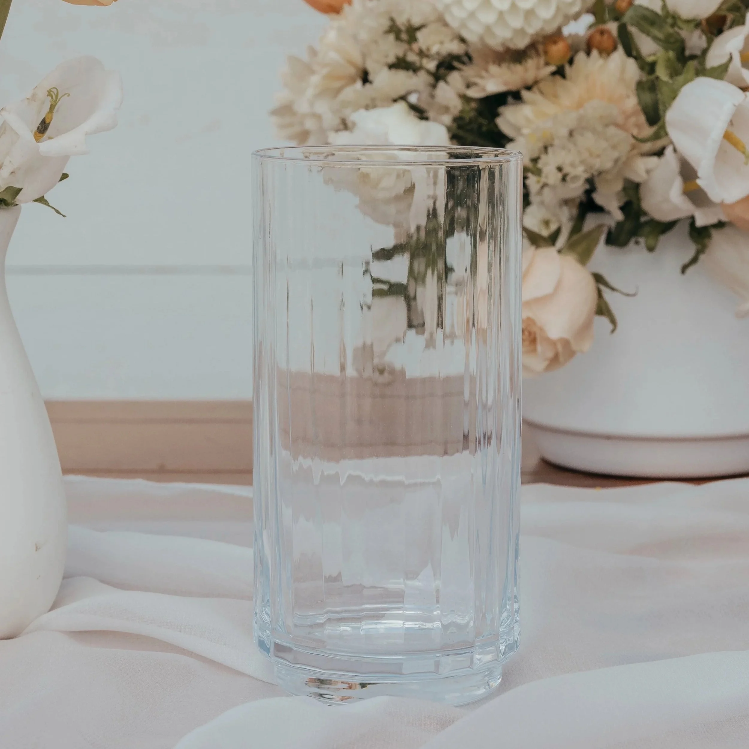 A clear, empty glass placed on a white cloth with a floral backdrop of white and beige flowers in vases.