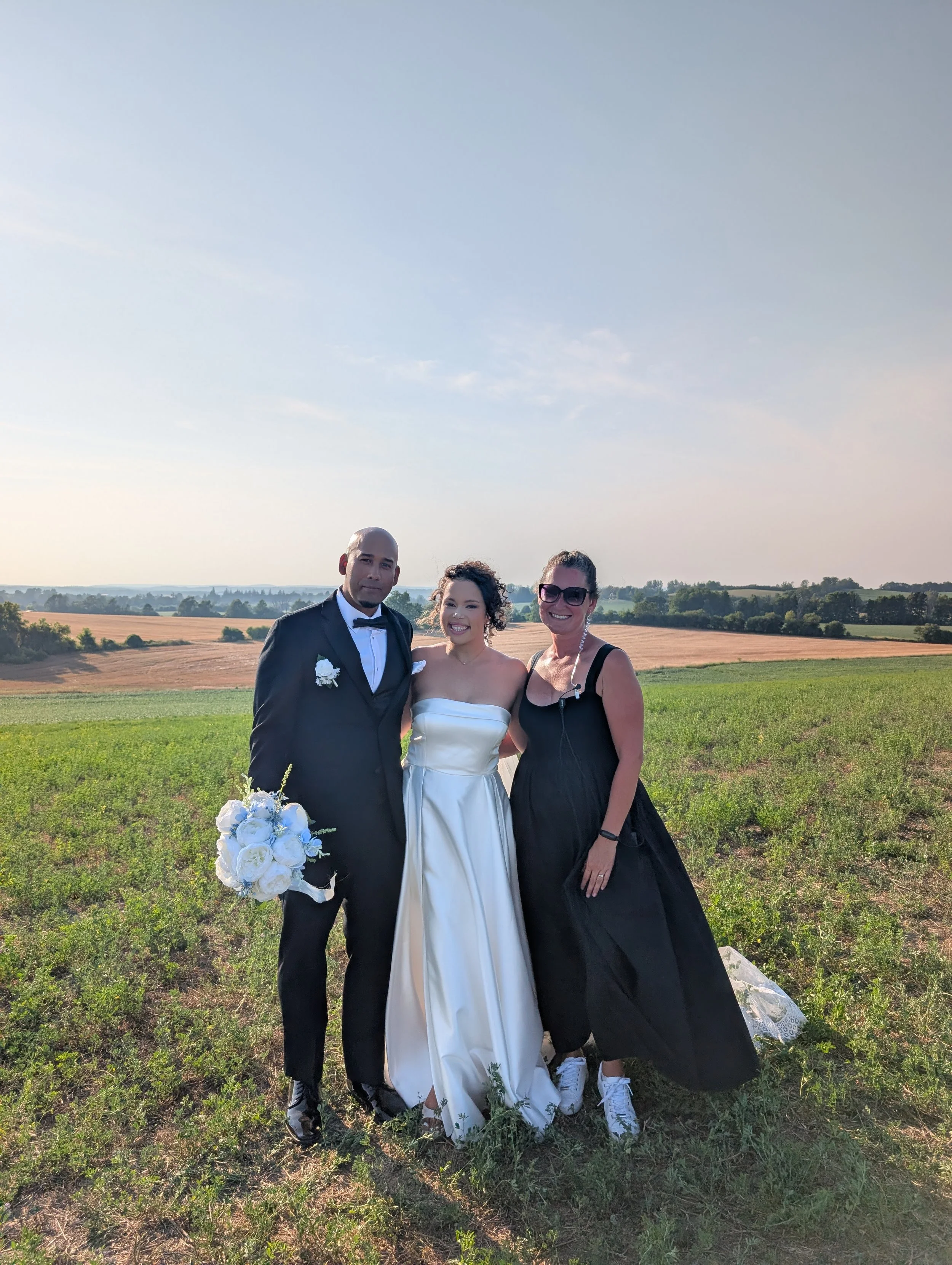 Three people stand together outdoors in a field, dressed in formal attire, with a man in a black tuxedo holding a bouquet of white and blue flowers, a woman in a white strapless wedding dress, and another woman in a black dress, smiling at the camera against a rural landscape background.
