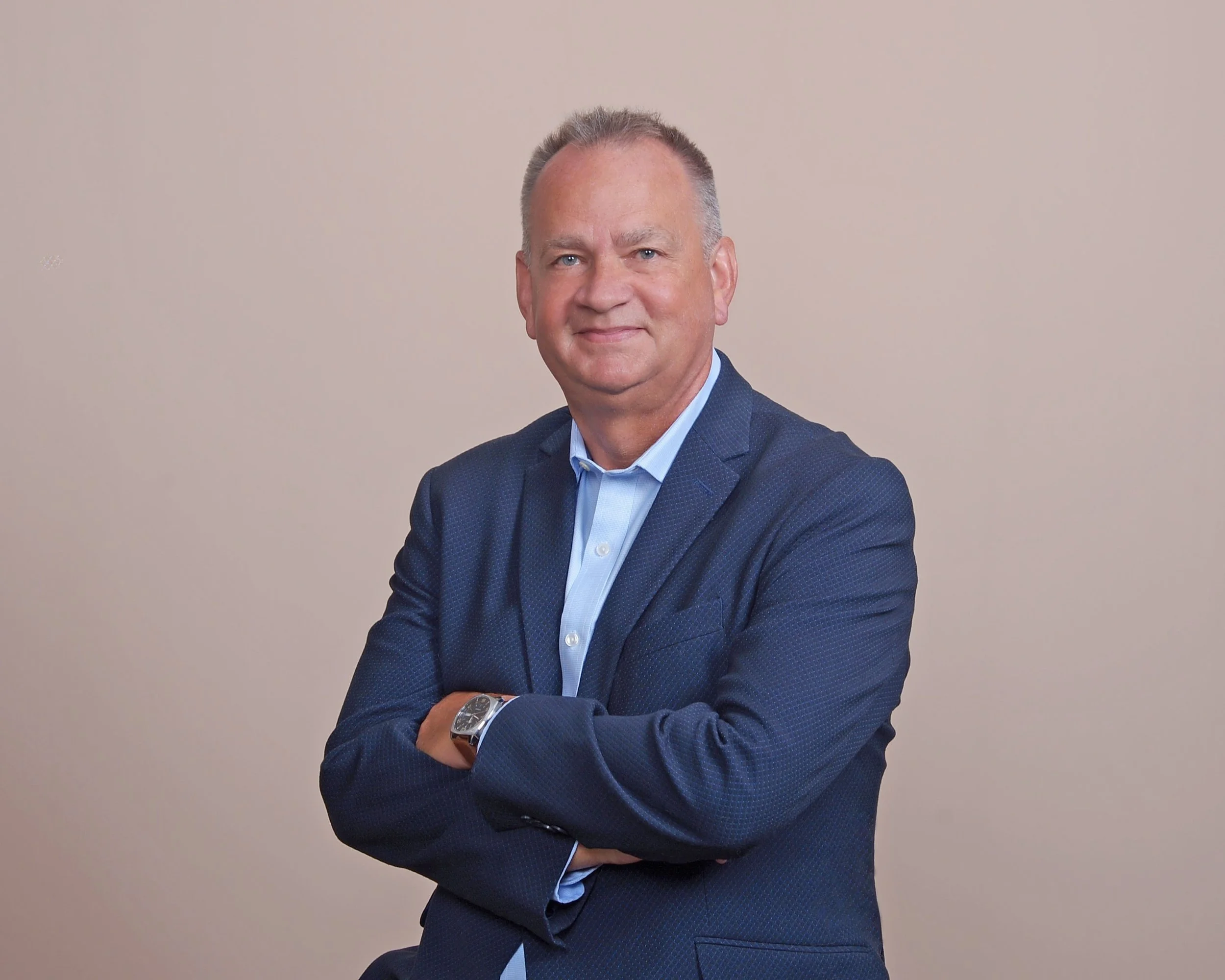 A middle-aged man in a blue suit with arms crossed, standing against a plain beige background.