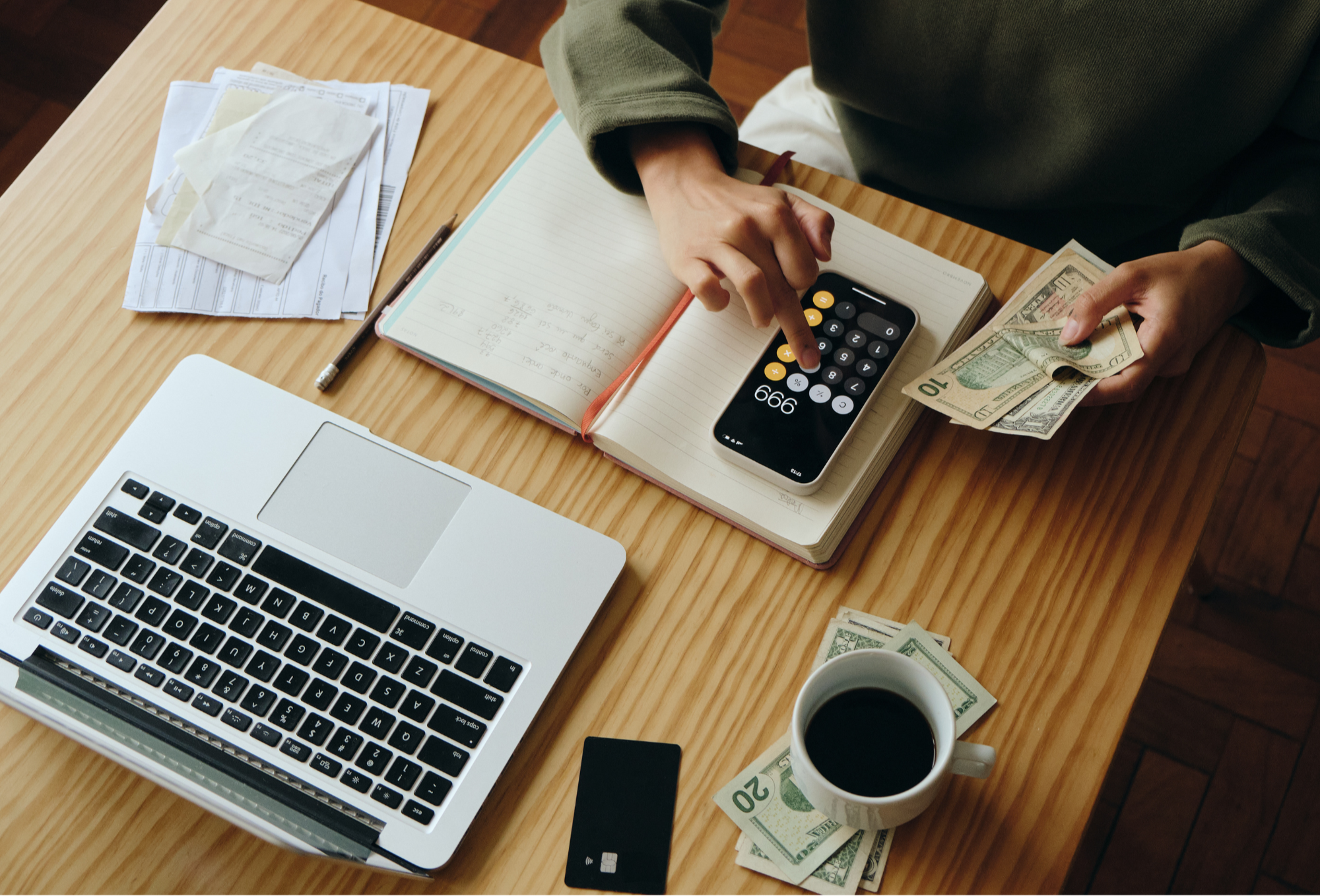 A person organizing cash, using a calculator and writing in a notebook on a wooden table with a laptop, coffee cup, debit card, and scattered receipts.