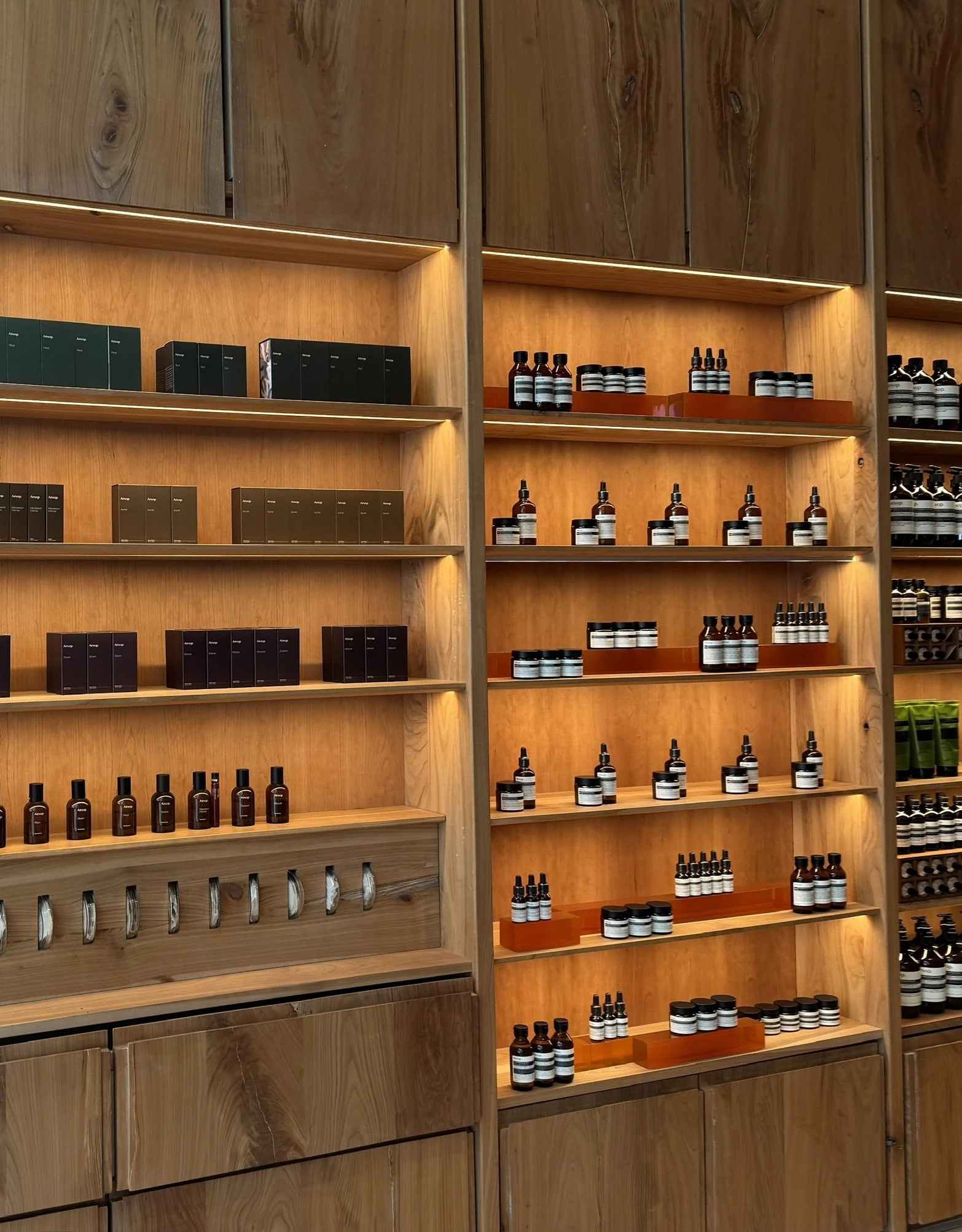 Wooden shelves displaying various skincare or cosmetic products in small bottles and jars, organized with some sections lit with warm lighting.