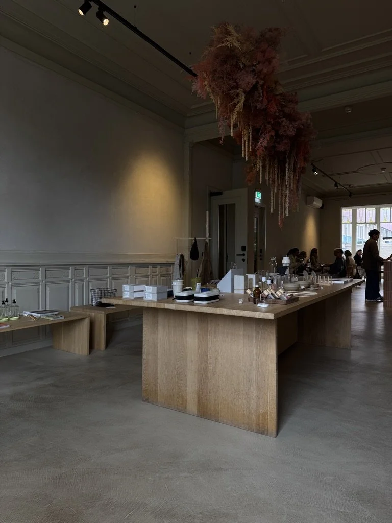 A retail display table in a store with various products and a woman browsing in the background, large dried flower arrangement hanging from the ceiling.