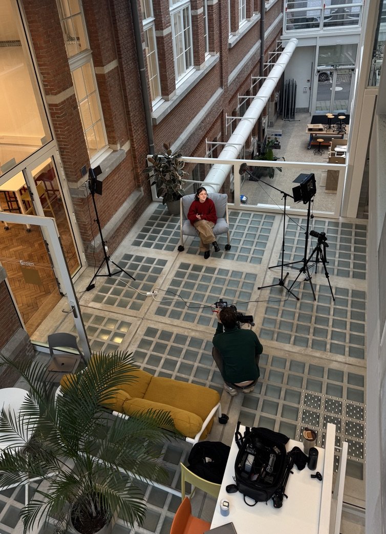 An indoor interview setup in a building atrium with a woman in a red sweater seated on a gray armchair, being filmed by a cameraman. Lighting equipment and a large potted plant are also visible.