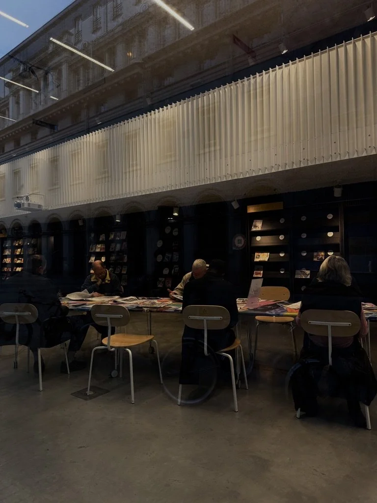 People sitting at tables, reading or looking at printed materials inside a bookstore or library with bookshelves in the background.