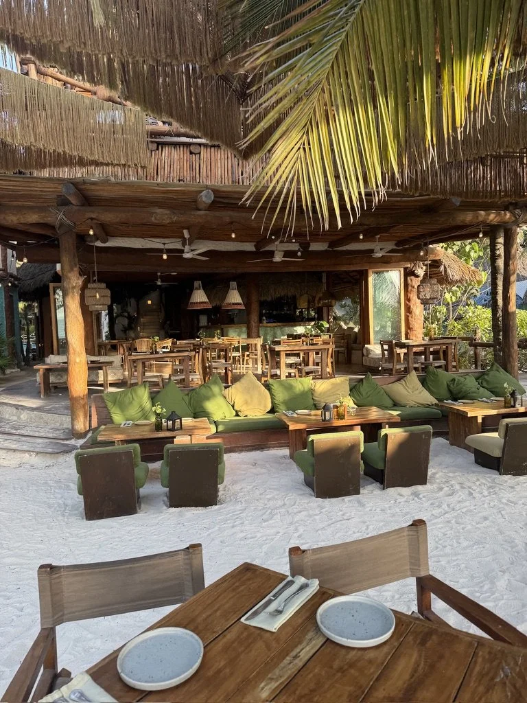 Beachside restaurant with wooden furniture, green and beige cushions, sand floor, thatched roof, and palm leaves overhead.
