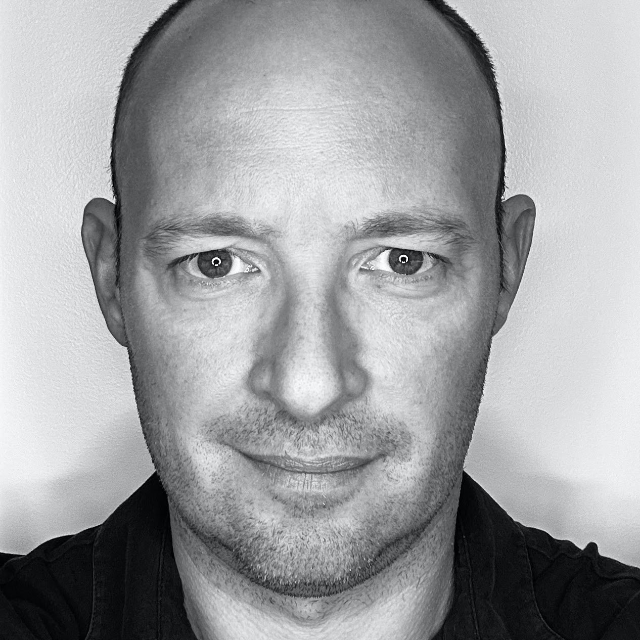 Close-up black-and-white portrait of a man with short hair, slight facial hair, and wearing a dark shirt, looking directly at the camera.
