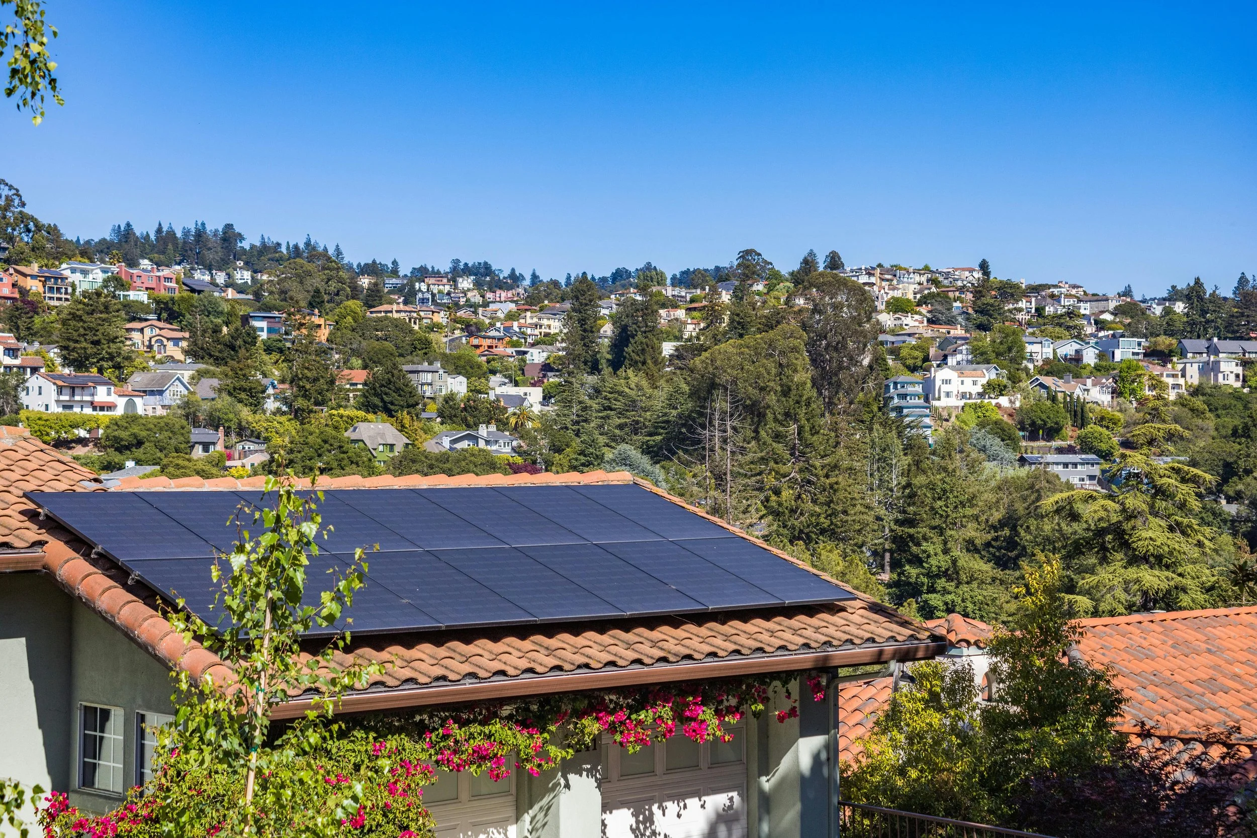 Häuser in einer hügeligen Stadt mit Solaranlagen auf dem Dach eines Hauses im Vordergrund, viele Bäume und ein blauer Himmel im Hintergrund.