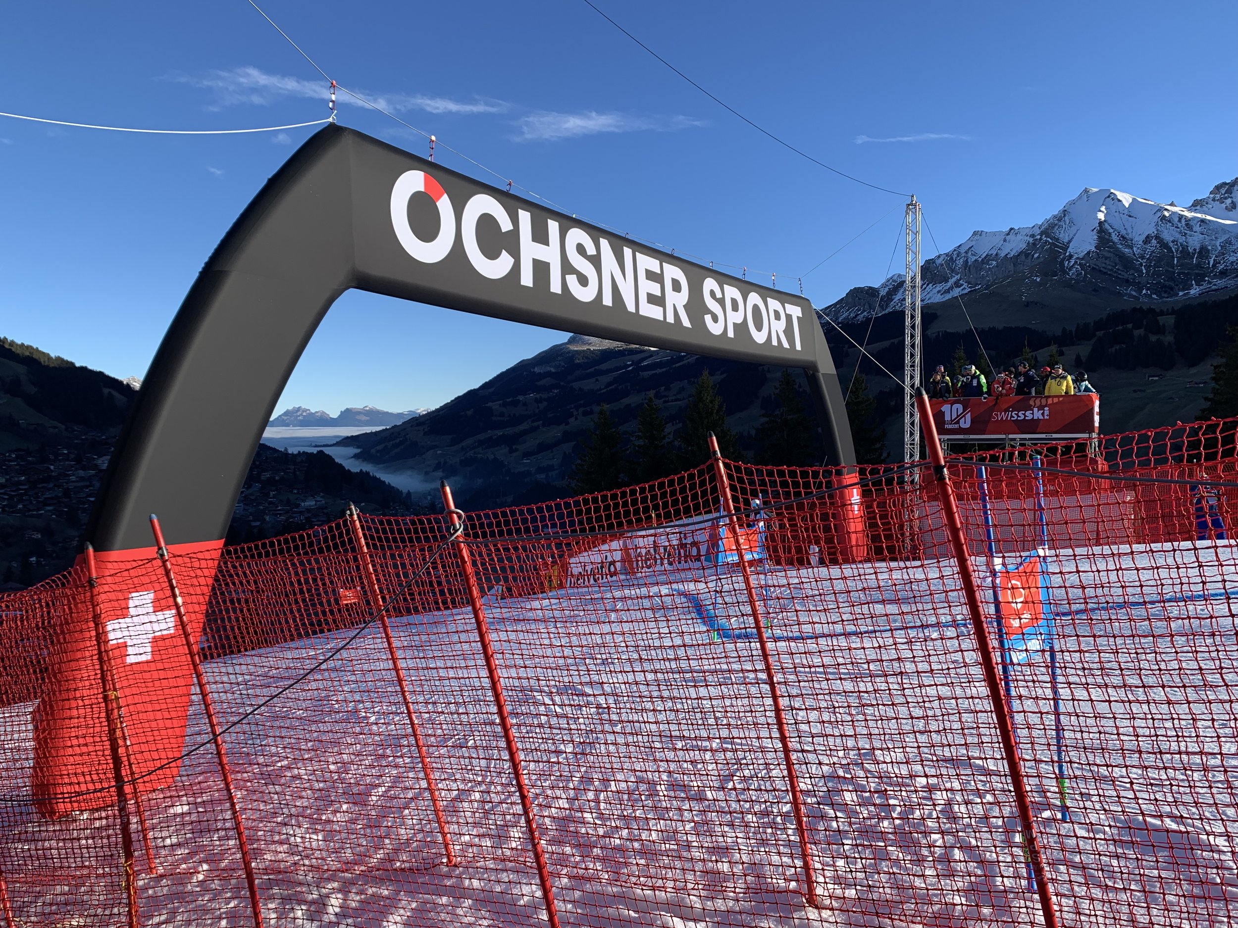 Aufblasbarer pneumatischer Torbogen für Ochsner Sport im schweizer Skiweltcup, roten Sicherheitszäunen und Gruppen von Skifahrern, mit schneebedeckten Bergen im Hintergrund.