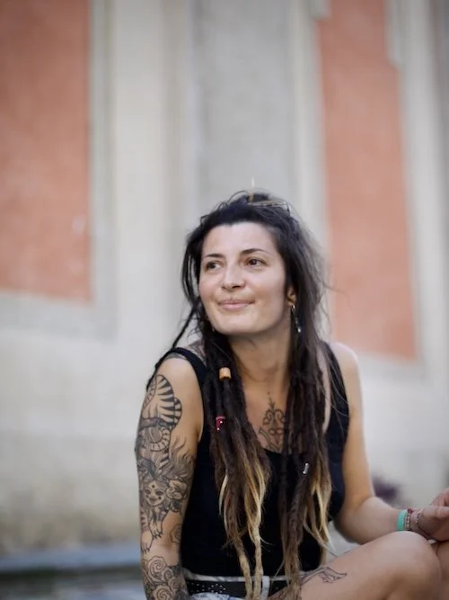 Jeune femme avec des tatouages, portant un débardeur noir, assise devant un mur en pierre, souriant légèrement.