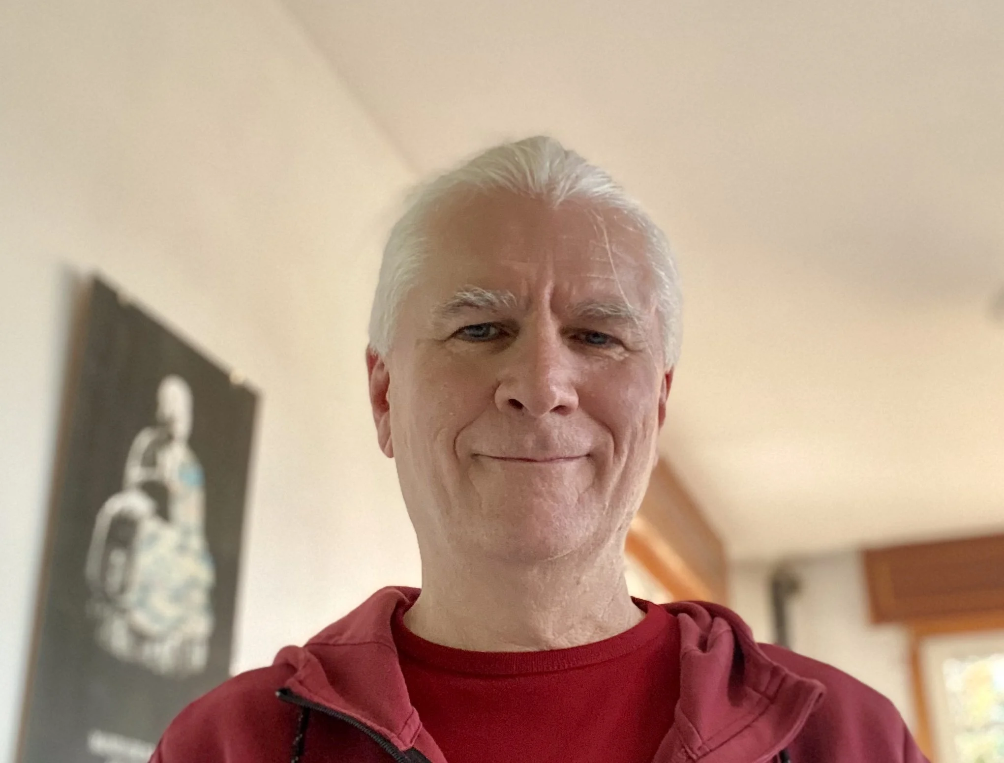 Hombre de cabello canoso y con expresión tranquila, usando sudadera roja, en un interior con paredes beige y ventana con cortinas de madera.