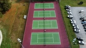 Four tennis courts with green playing surfaces and red surrounding area, viewed from above, with parked cars and grassy areas nearby.