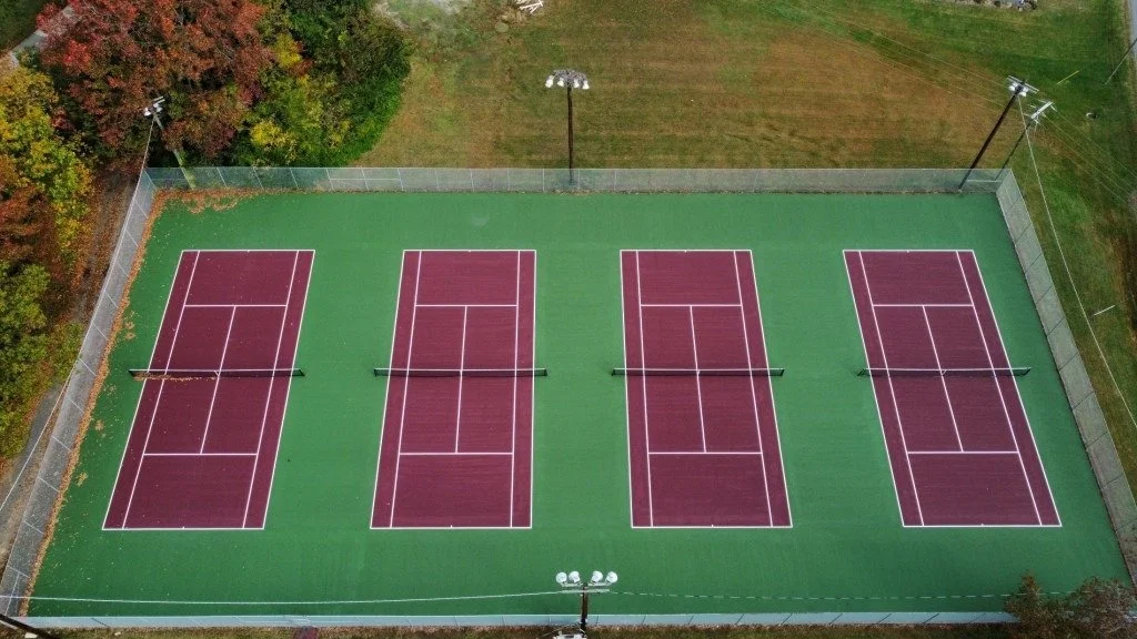 Four tennis courts with purple surfaces and white lines, surrounded by a chain-link fence, situated in a grassy area with trees nearby.