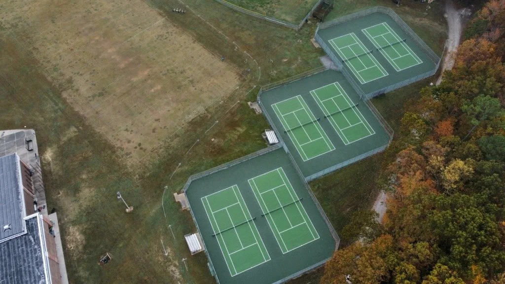 Three outdoor tennis courts with green surfaces, white lines, and fencing, surrounded by trees and a grassy area.