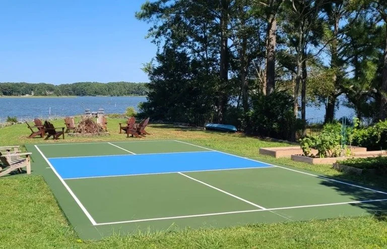 Tennis court by a lake, with Adirondack chairs and a fire pit in the background, surrounded by trees.