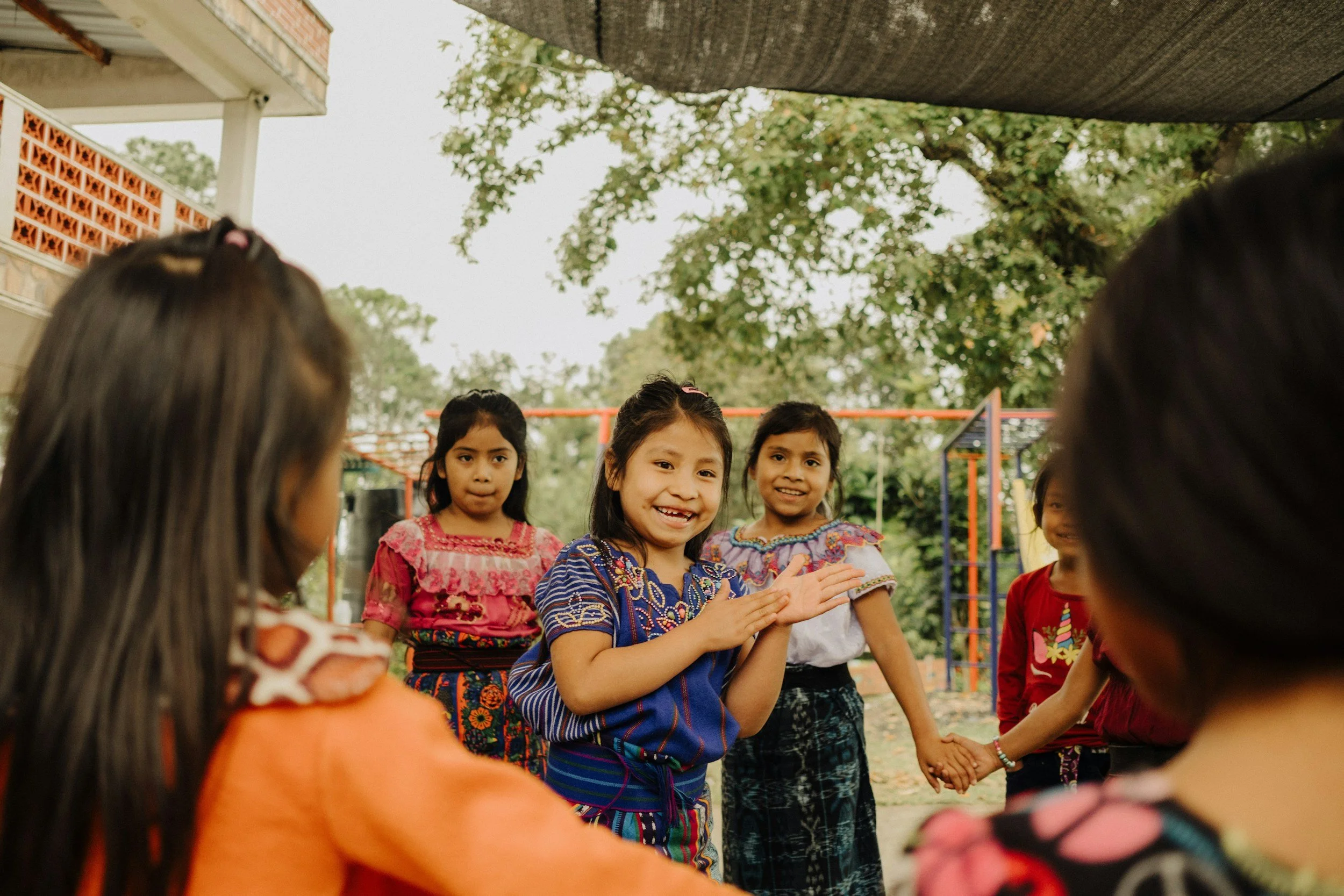 A group of young girls from different backgrounds holding hands and smiling, standing outside under a shaded area, with playground equipment and trees in the background.