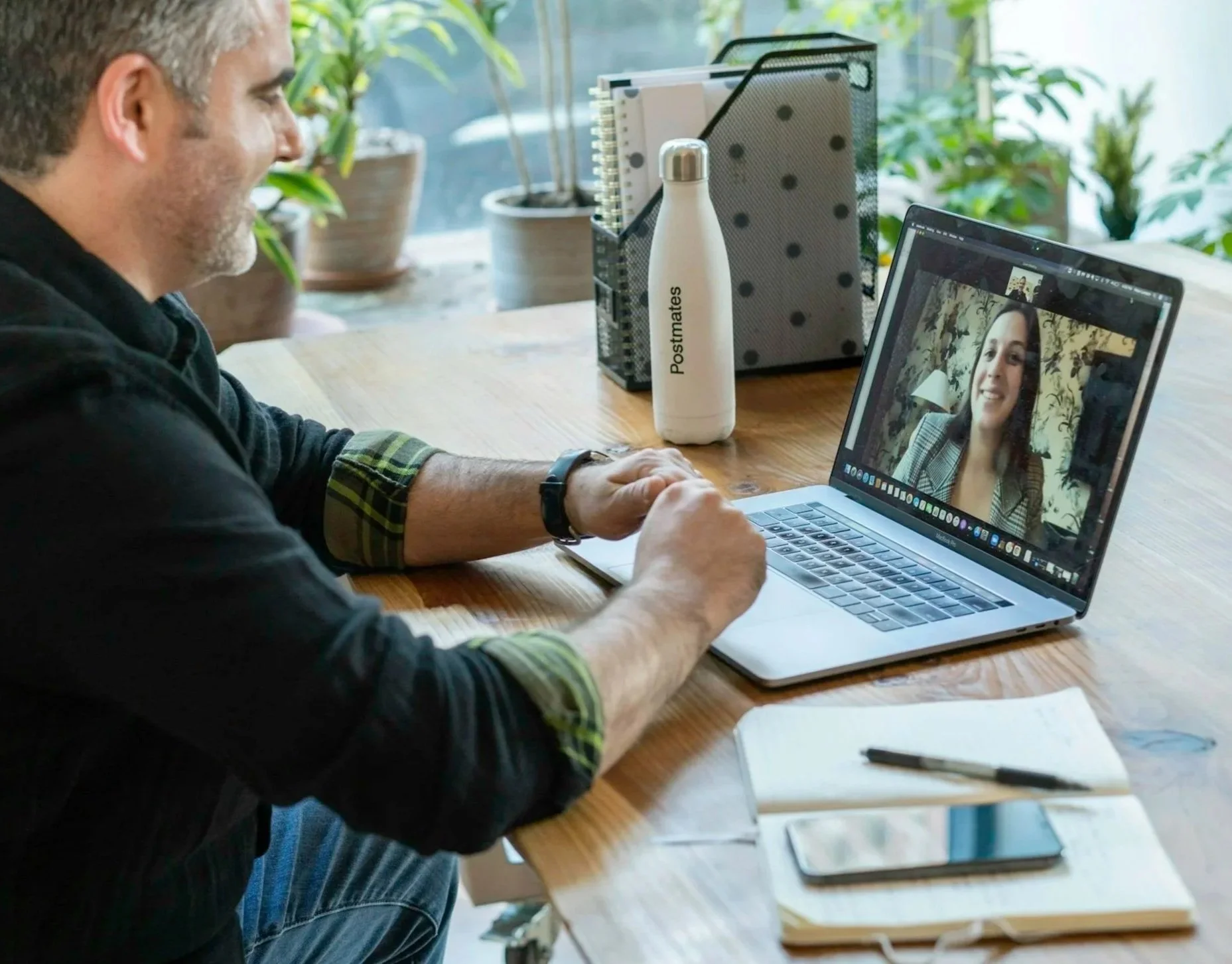 A man sitting at a desk on a video call with a woman on his laptop, surrounded by notebooks, a pen, a smartphone, plants, and office supplies.