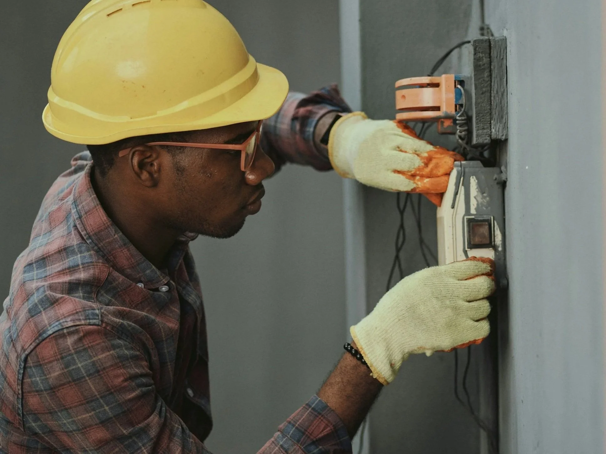 A man wearing a yellow hard hat, glasses, gloves, and a plaid shirt working on an electrical outlet on a wall.