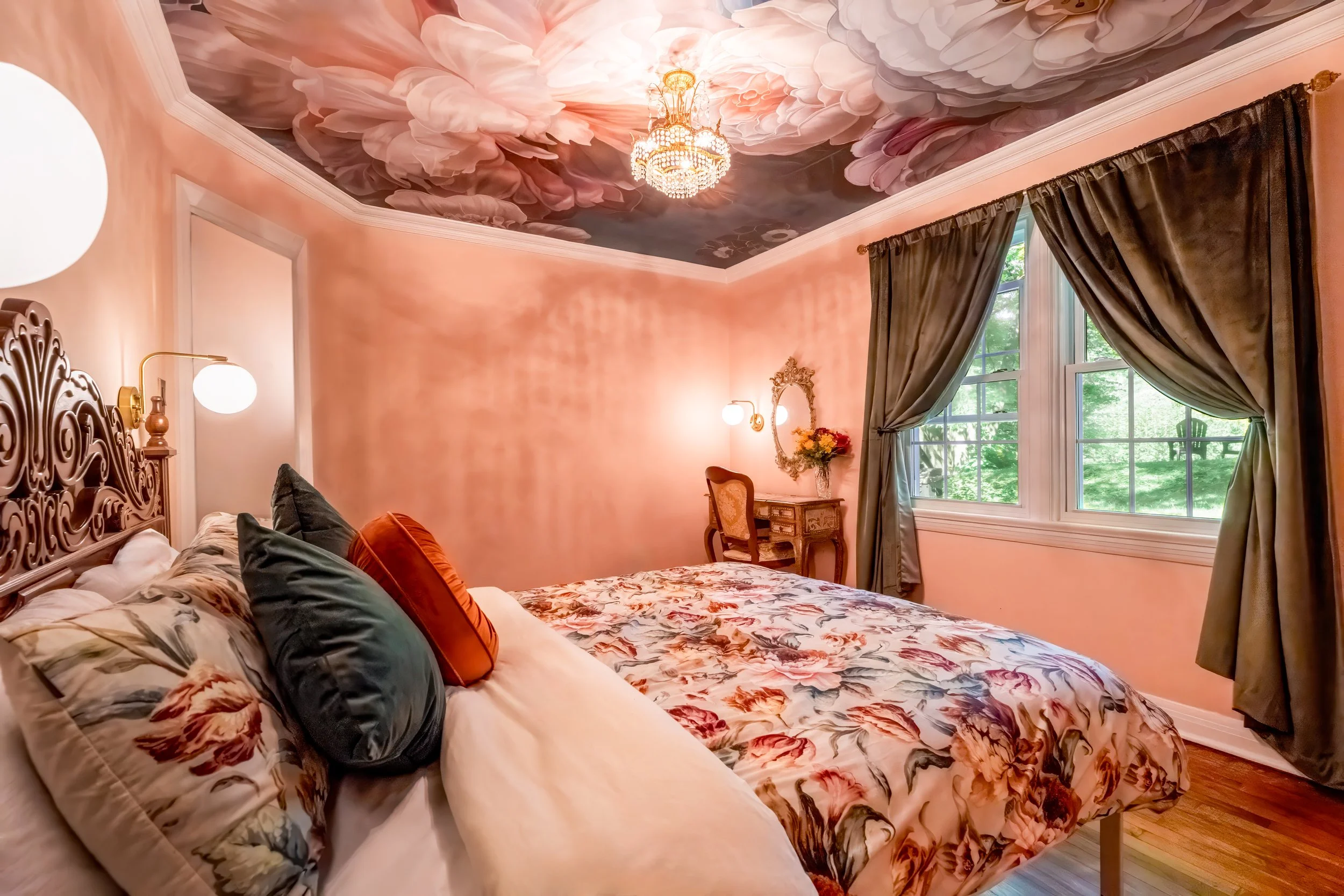 A bedroom with pink walls and floral bedding, ornate wooden headboard, green and orange pillows, large window with green curtains, vintage desk with flowers, mirror, floral ceiling mural, chandelier, and warm lighting.