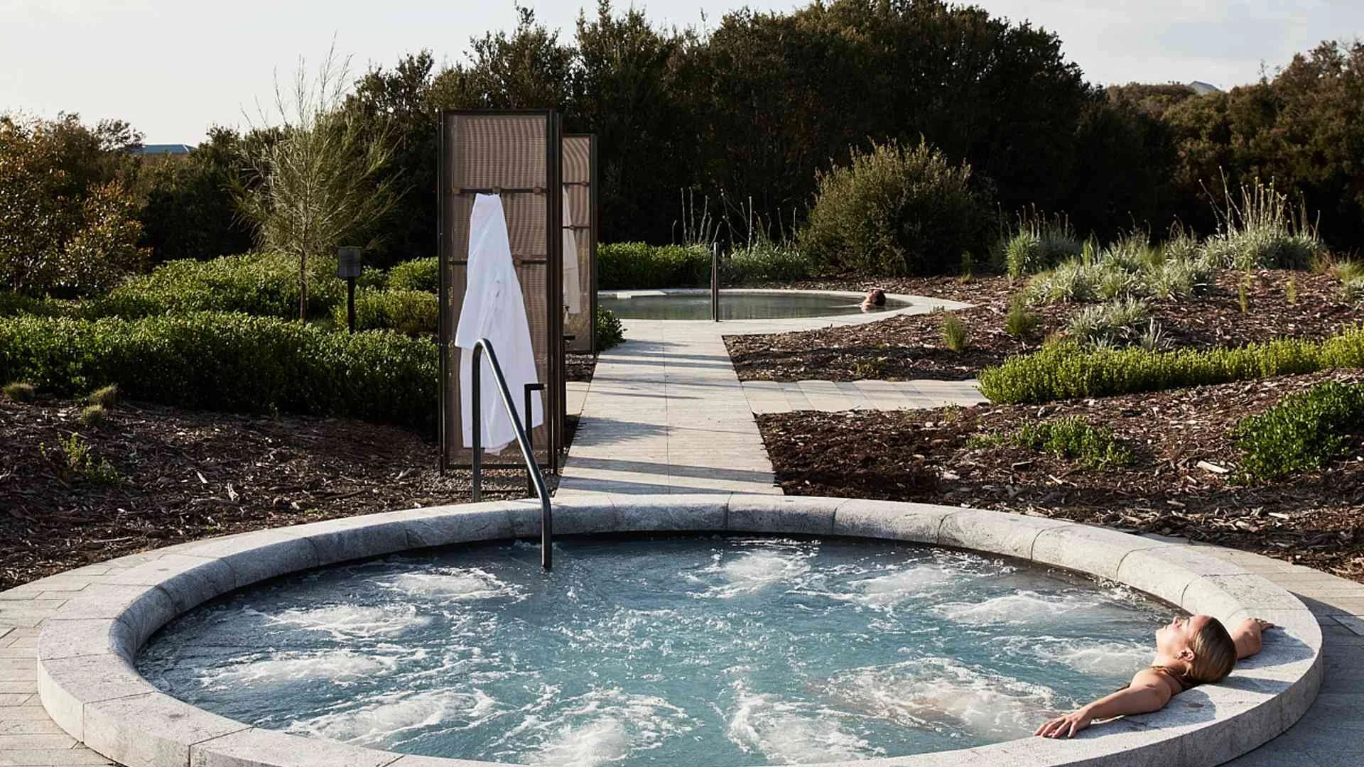 Person relaxing in hot tub outdoors near a pathway leading to a small pool, surrounded by landscaped bushes and trees.