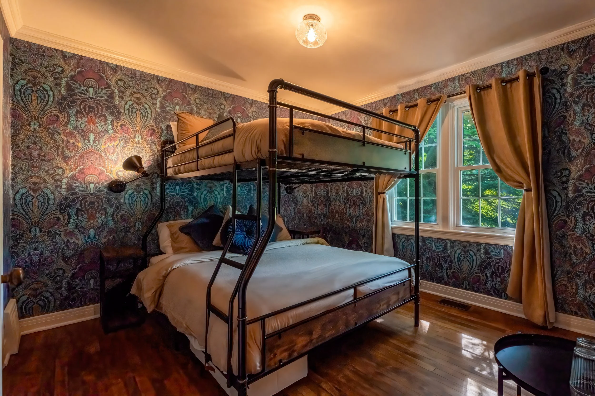 A bedroom with a loft bed over a double bed, featuring dark patterned wallpaper, wooden flooring, two windows with curtains, and a black wall-mounted reading lamp.