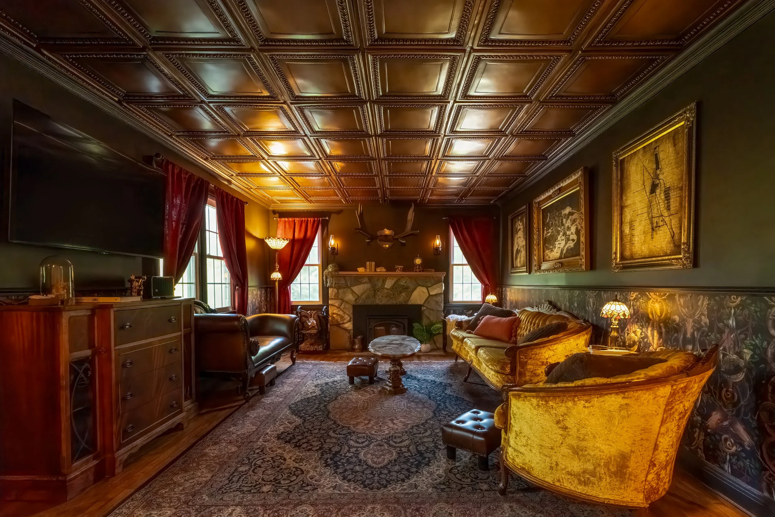 Vintage living room with dark green walls, ornate wood ceiling, red curtains, antique furniture including two yellow velvet armchairs, a green velvet couch, a wooden cabinet with a TV on top, and decorated with artwork, lamps, and a fireplace.