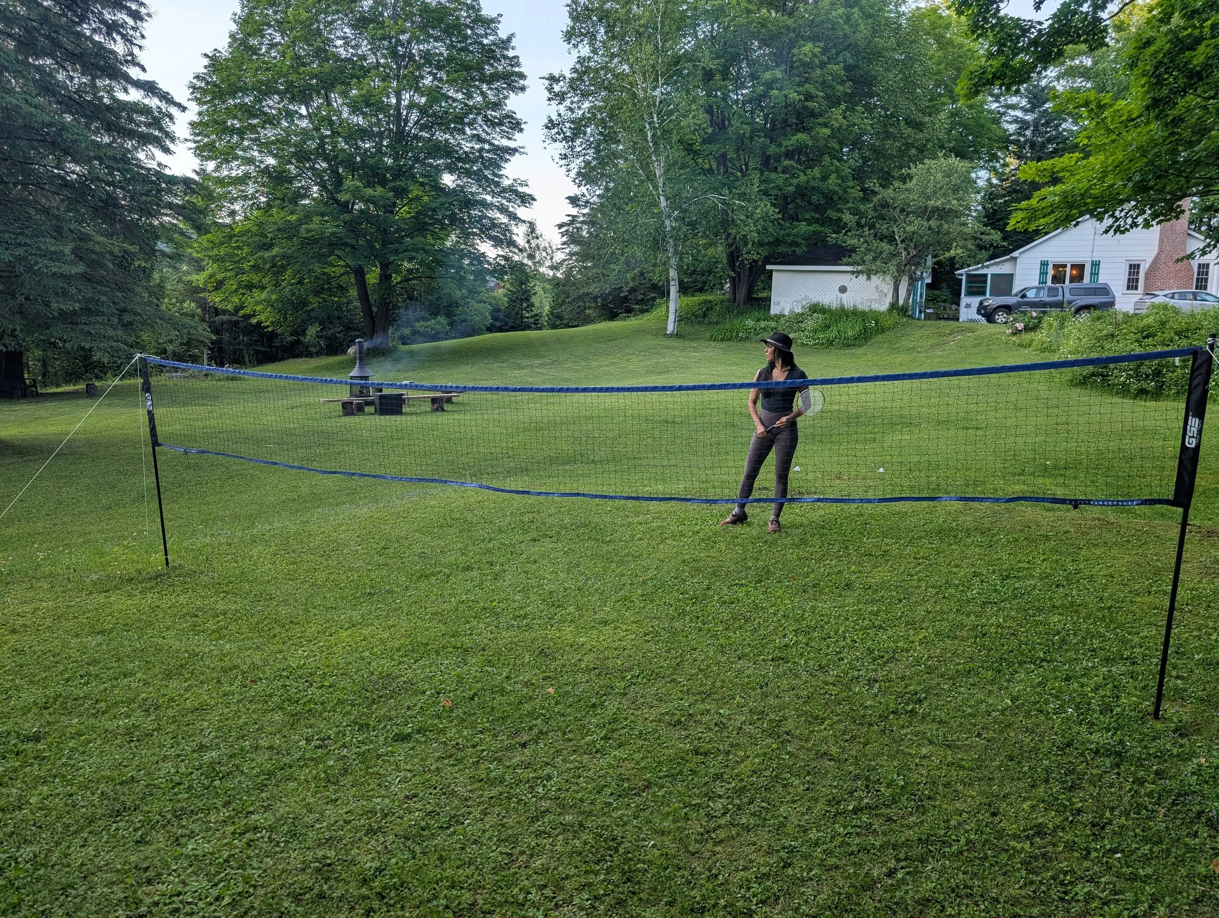 A woman wearing a black hat, black shirt, and gray pants stands on a grassy lawn holding a badminton racket beside a badminton net set up outdoors. In the background, there are trees, a small white building, a stone fire pit with benches, and a white
