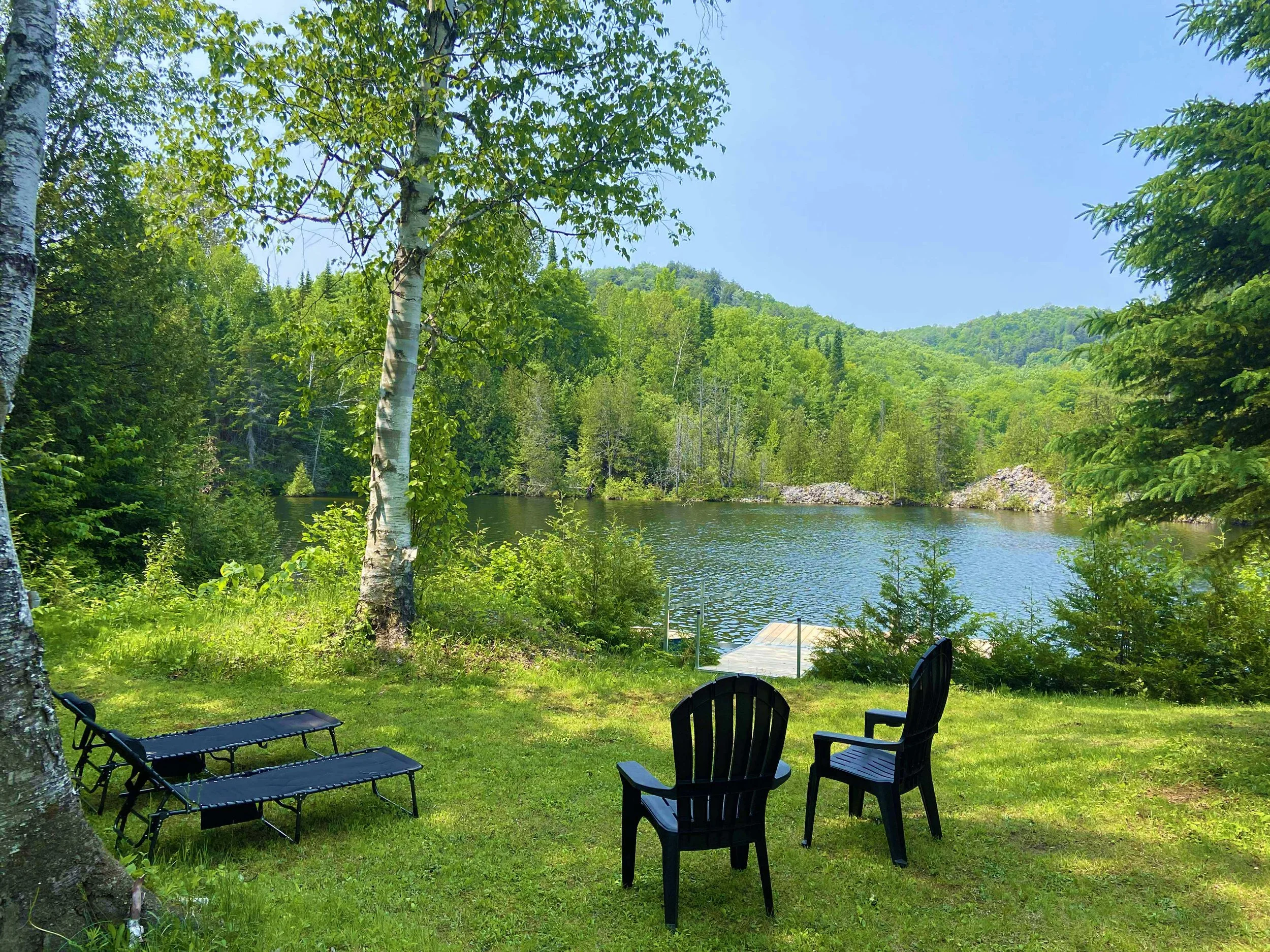 A peaceful lakeside scene with two black Adirondack chairs and two black lounge chairs on a grassy area, surrounded by trees and hills under a clear blue sky.
