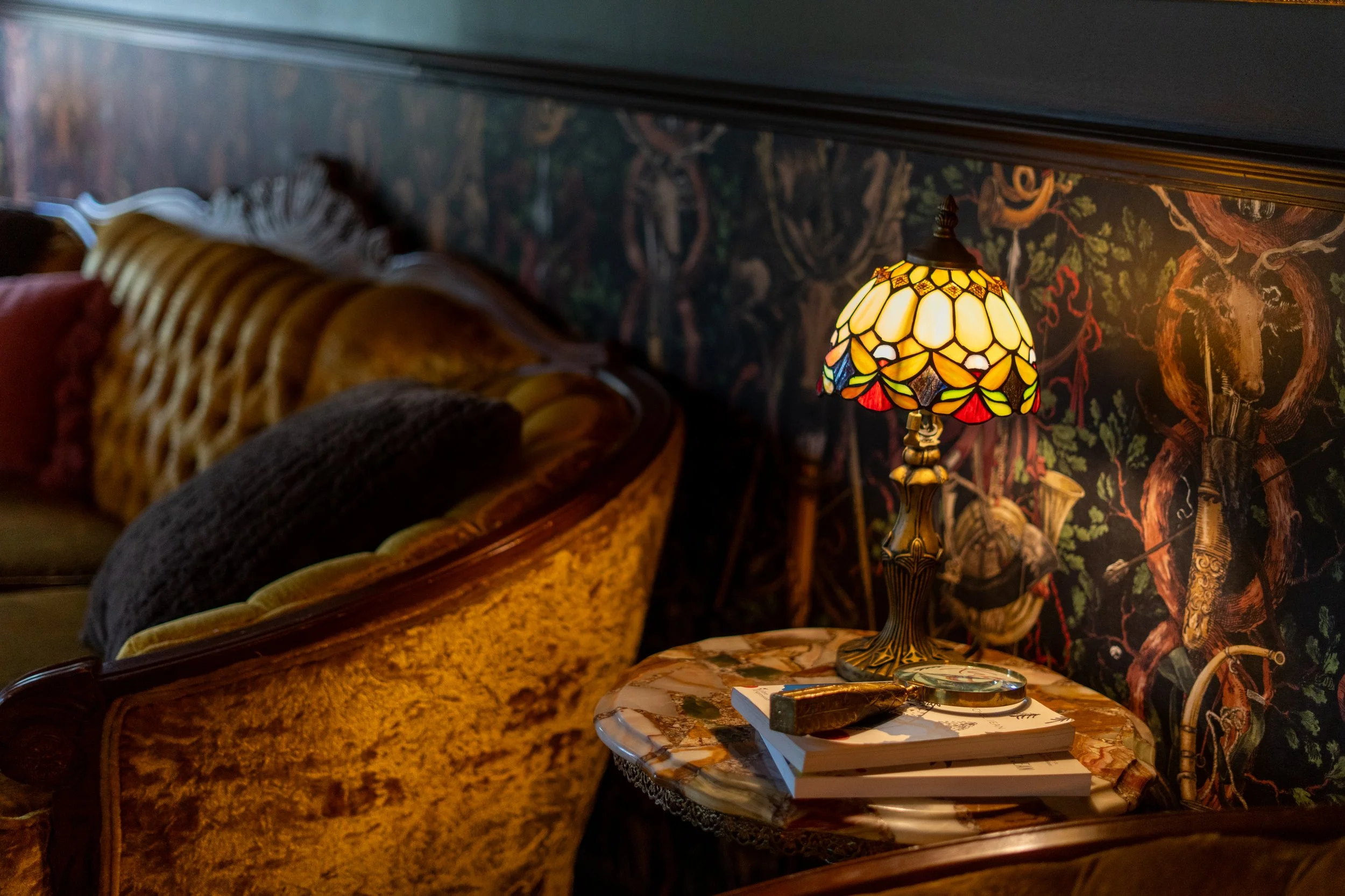 A cozy living room corner featuring a vintage sofa with cushions, a marble-topped side table with stacked books, a Tiffany-style stained glass table lamp, and a dark ornate wallpaper with botanical and animal motifs.