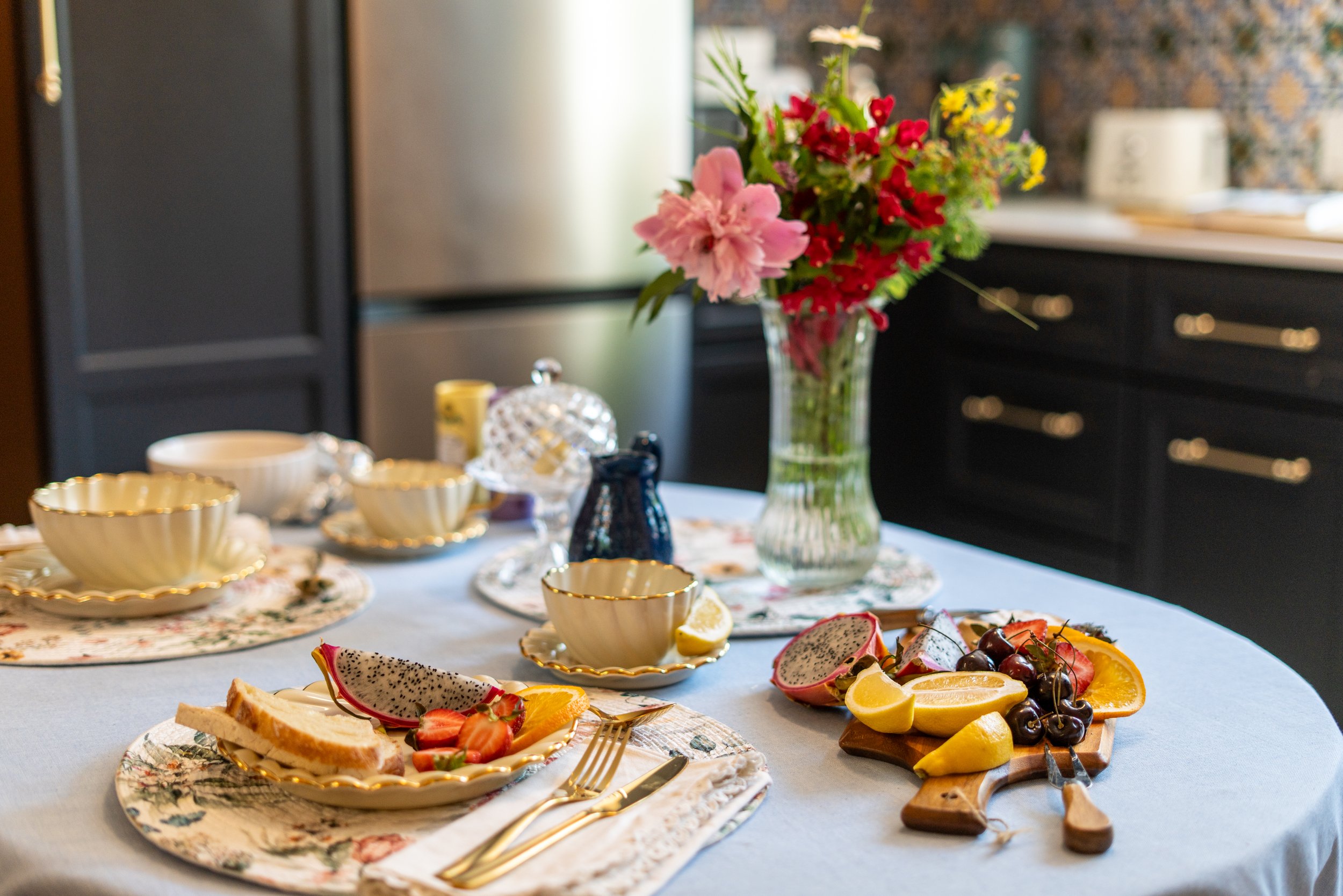 A table set for a meal with dessert and fruit, including a large vase of pink and red flowers as a centerpiece.