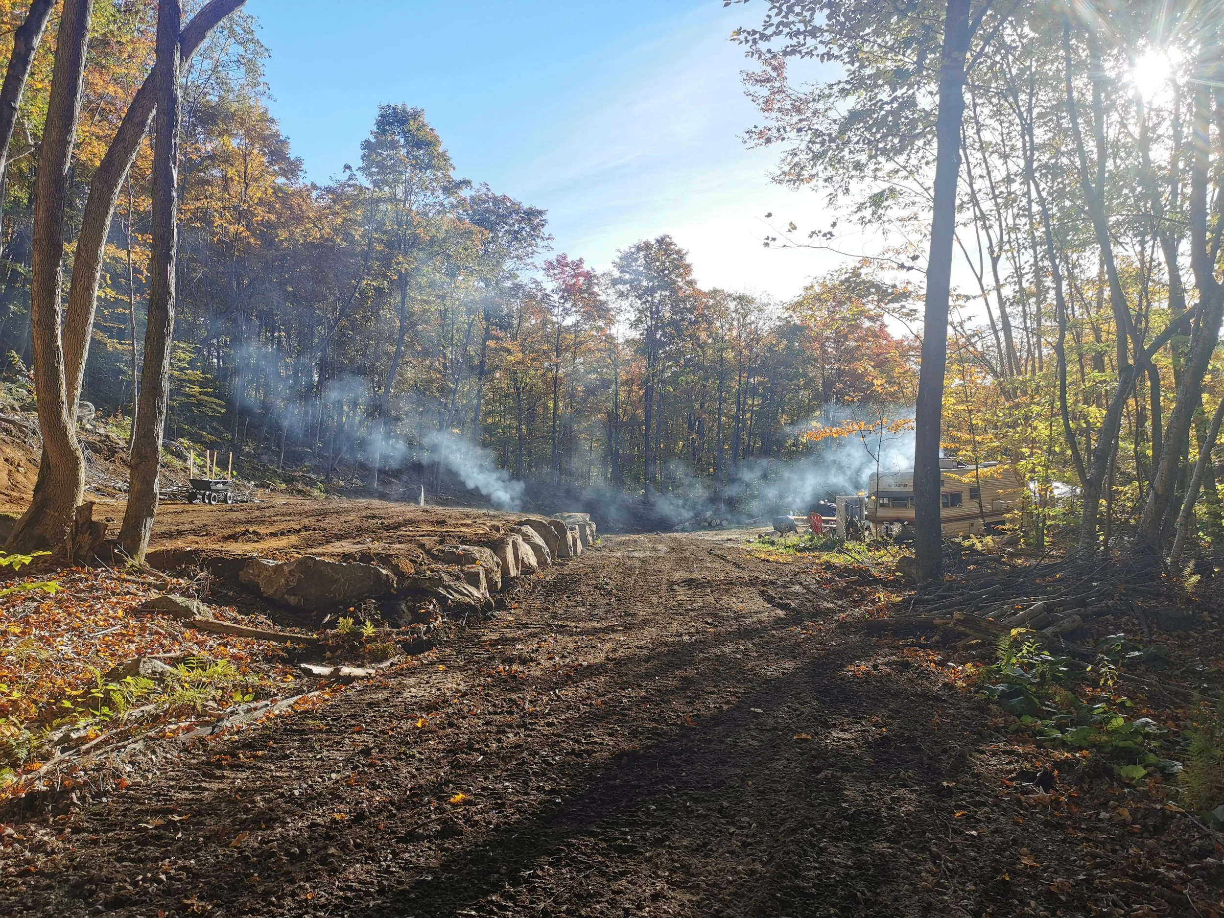 A dirt road in a forest with smoke rising from fires in the background, trees with autumn leaves, and sunlight filtering through the trees.