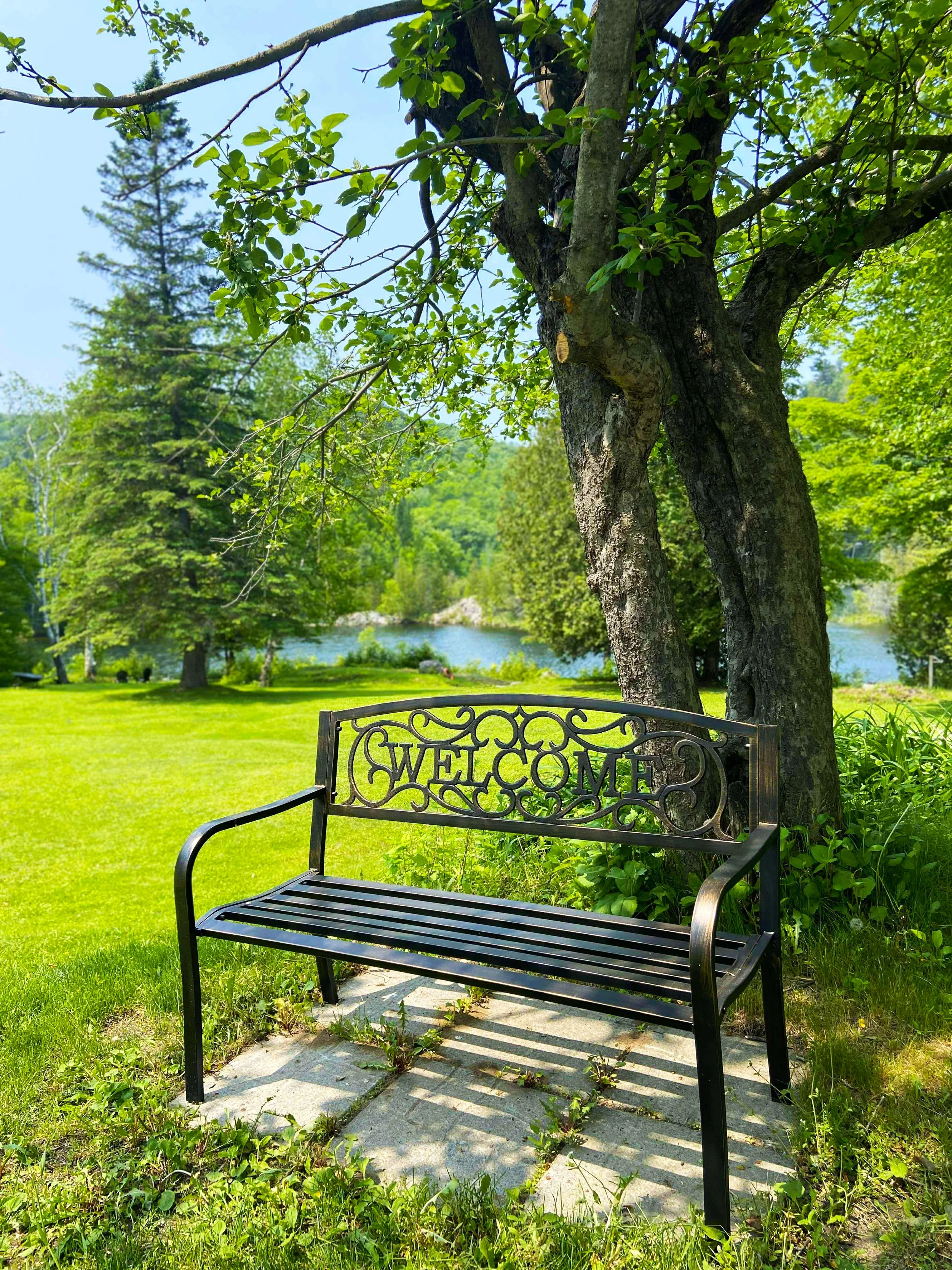 A black metal bench with a decorative backrest spelling 'WELCOME' sits on a stone patio in a lush green park near a tree. In the background, there is a river, trees, and a grassy area under a bright blue sky.