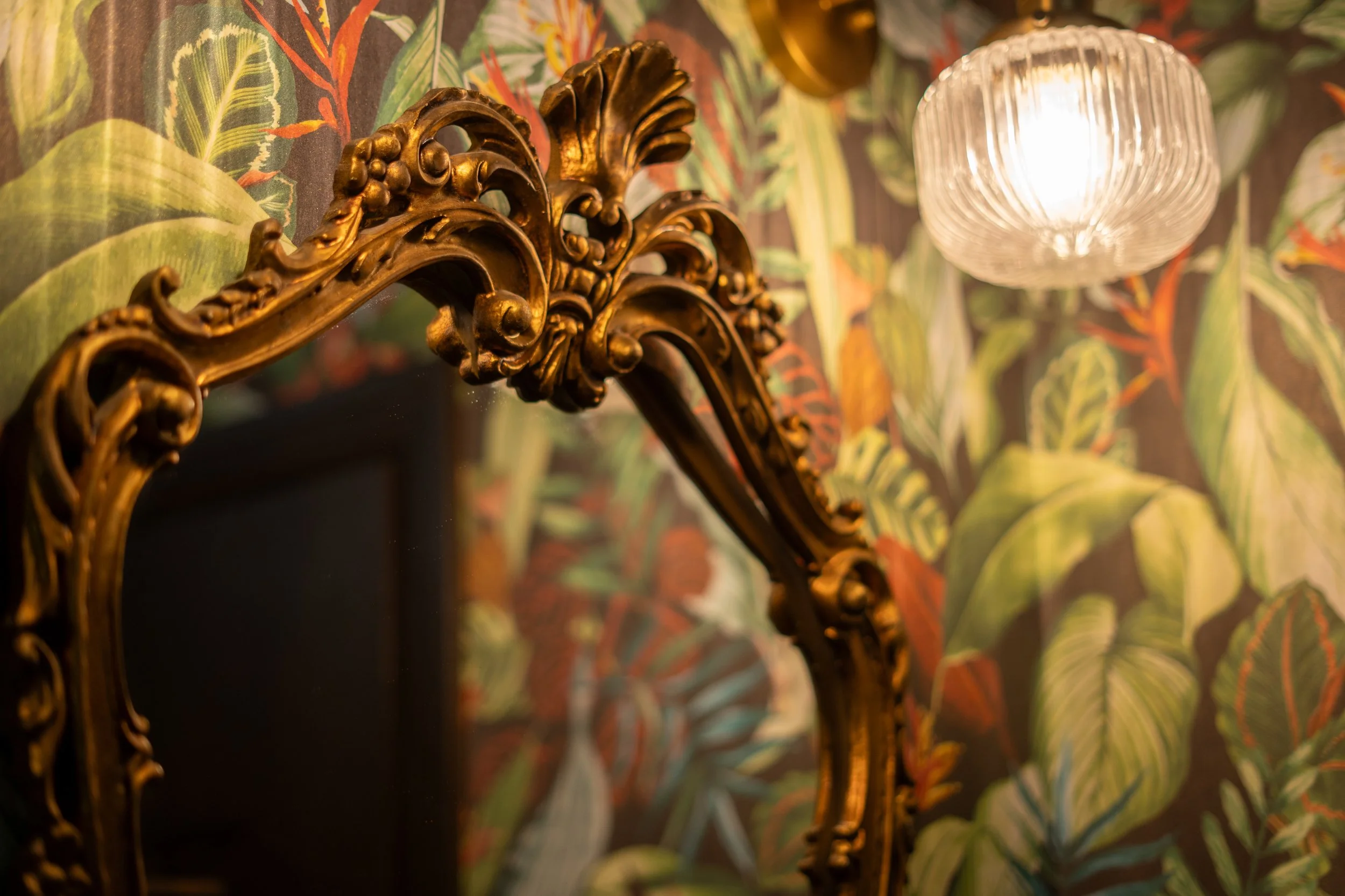 Close-up of an ornate gold-framed mirror against a wall with tropical leaf-patterned wallpaper, and a glass pendant light hanging above.