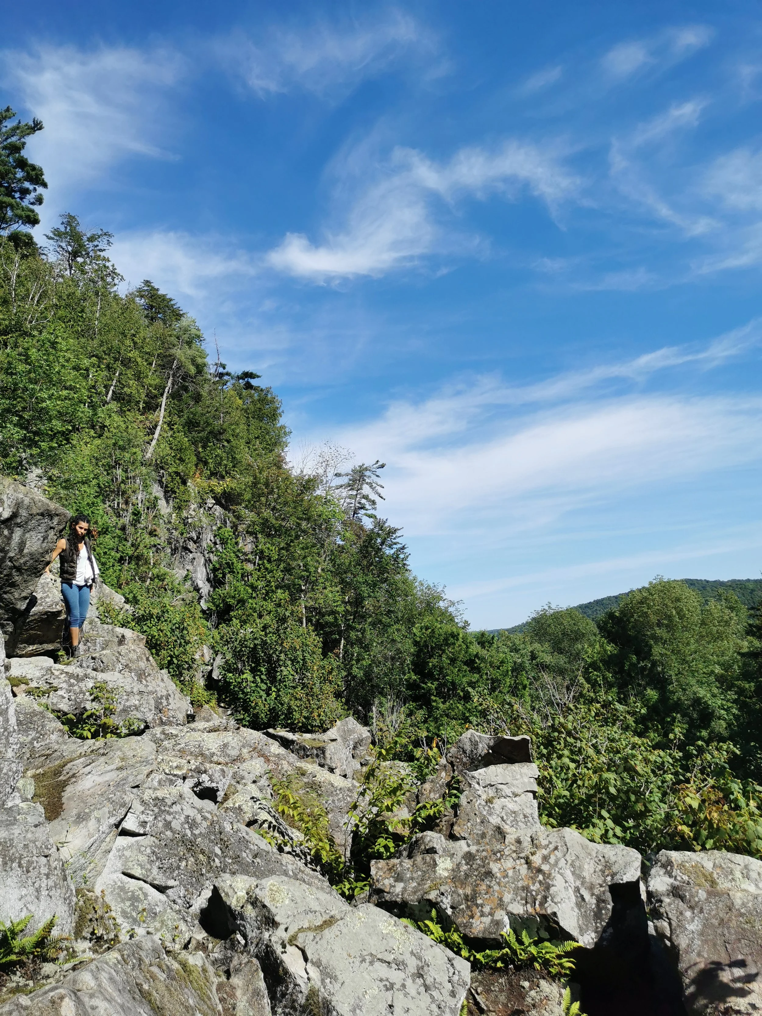 Woman hiking down rocky terrain with lush green trees and a blue sky with wispy clouds in the background.