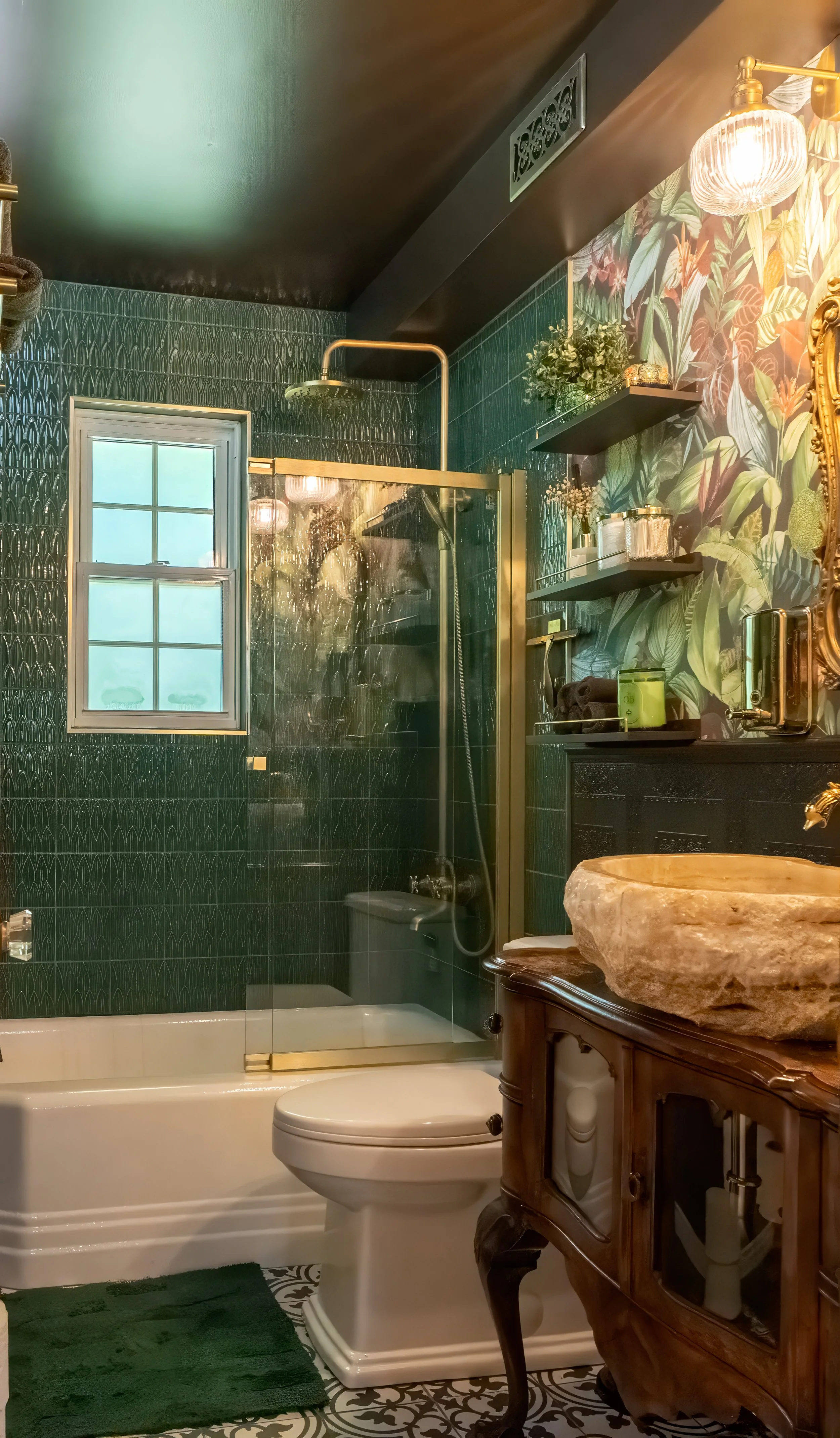 A bathroom with dark green textured wall tiles, a small window, a glass shower enclosure with brass trim, a white bathtub, a vintage-style toilet, a marble vessel sink on a wooden vanity, black wall-mounted shelves with decorative items, a gold-frame