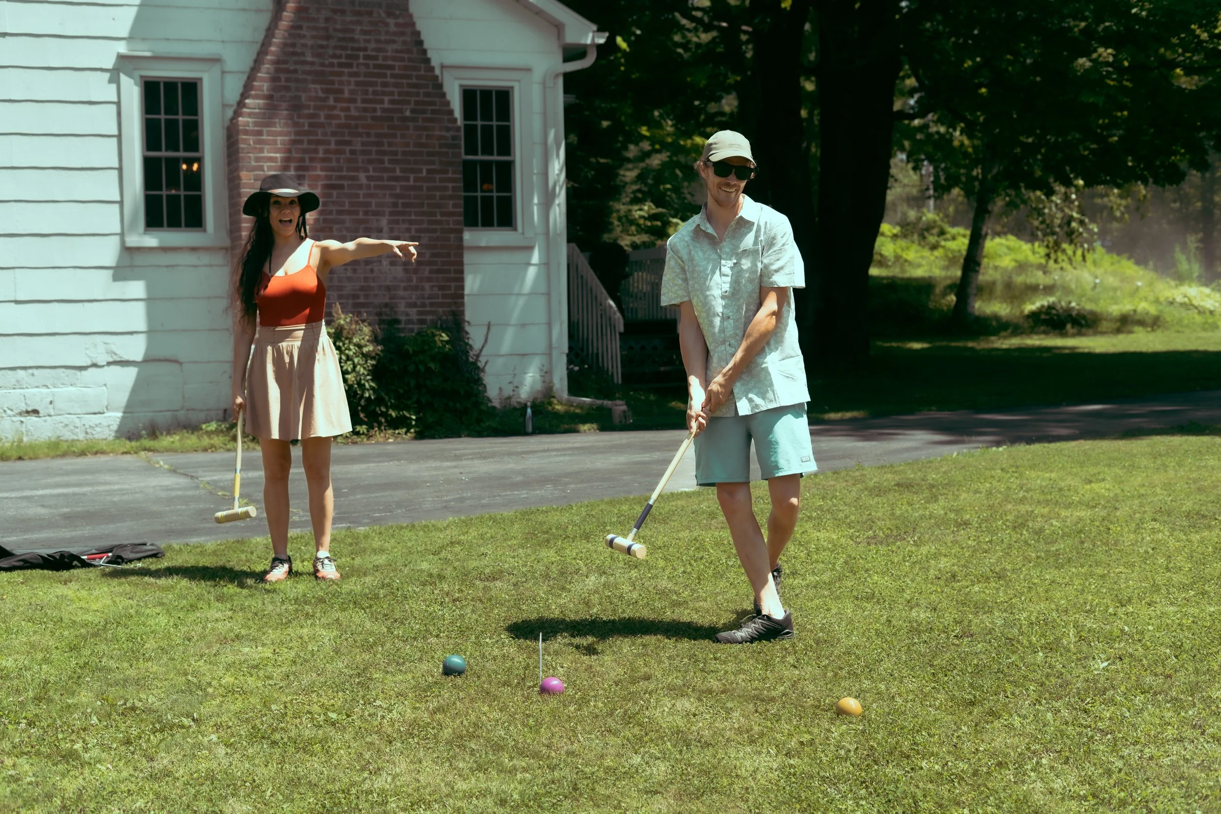 Two young adults playing croquet outdoors on a sunny day near a white house with red brick accents. The woman is wearing a red top, beige skirt, and black hat, pointing while smiling. The man is in a light-colored shirt, shorts, and sunglasses, holdi