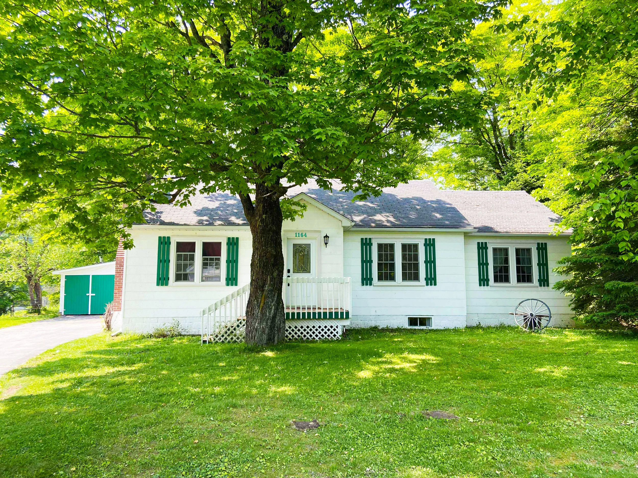 White house with green shutters and a front porch, surrounded by trees and green grass.