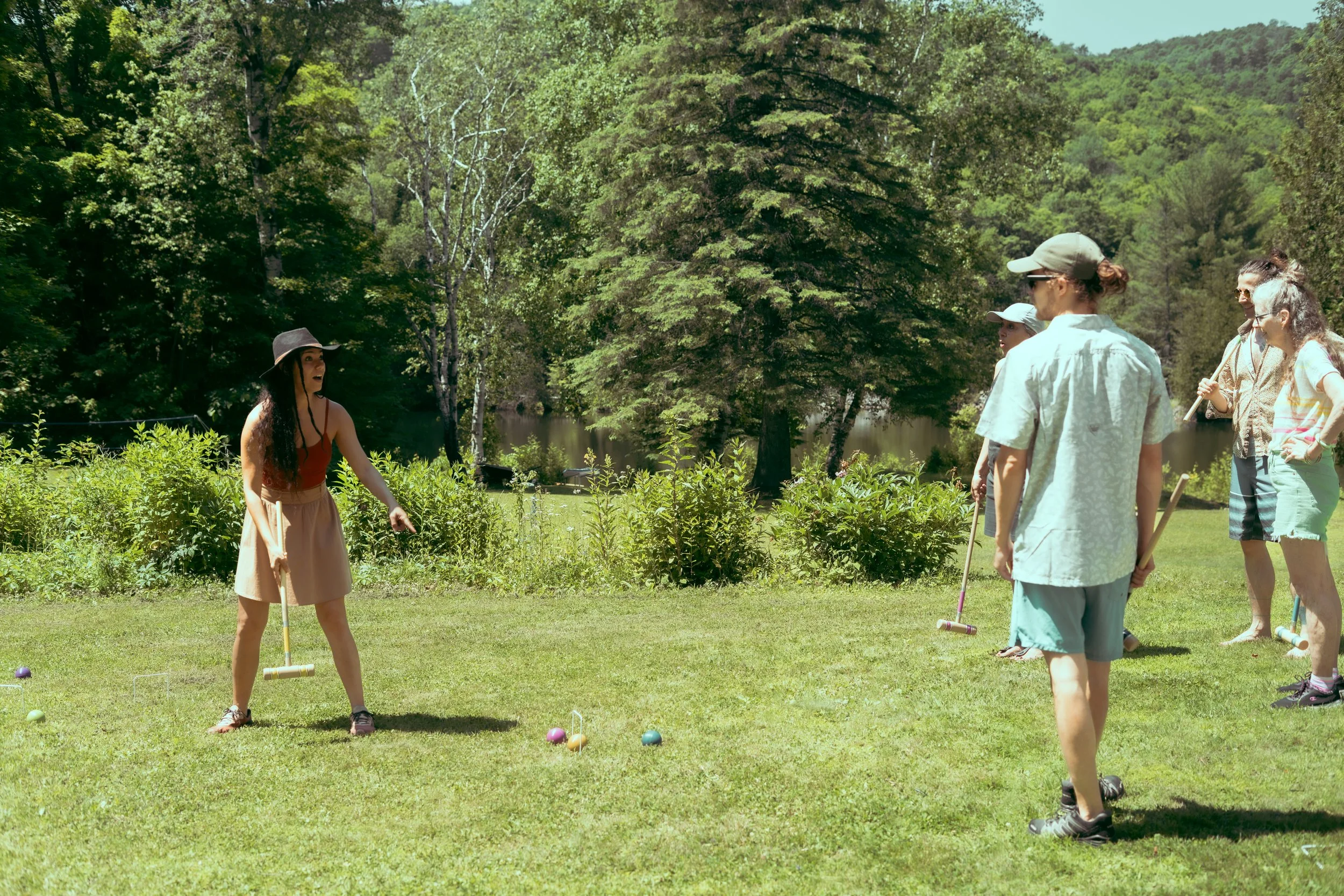 A group of five women playing croquet outdoors on grass near a lake, surrounded by trees and hills.