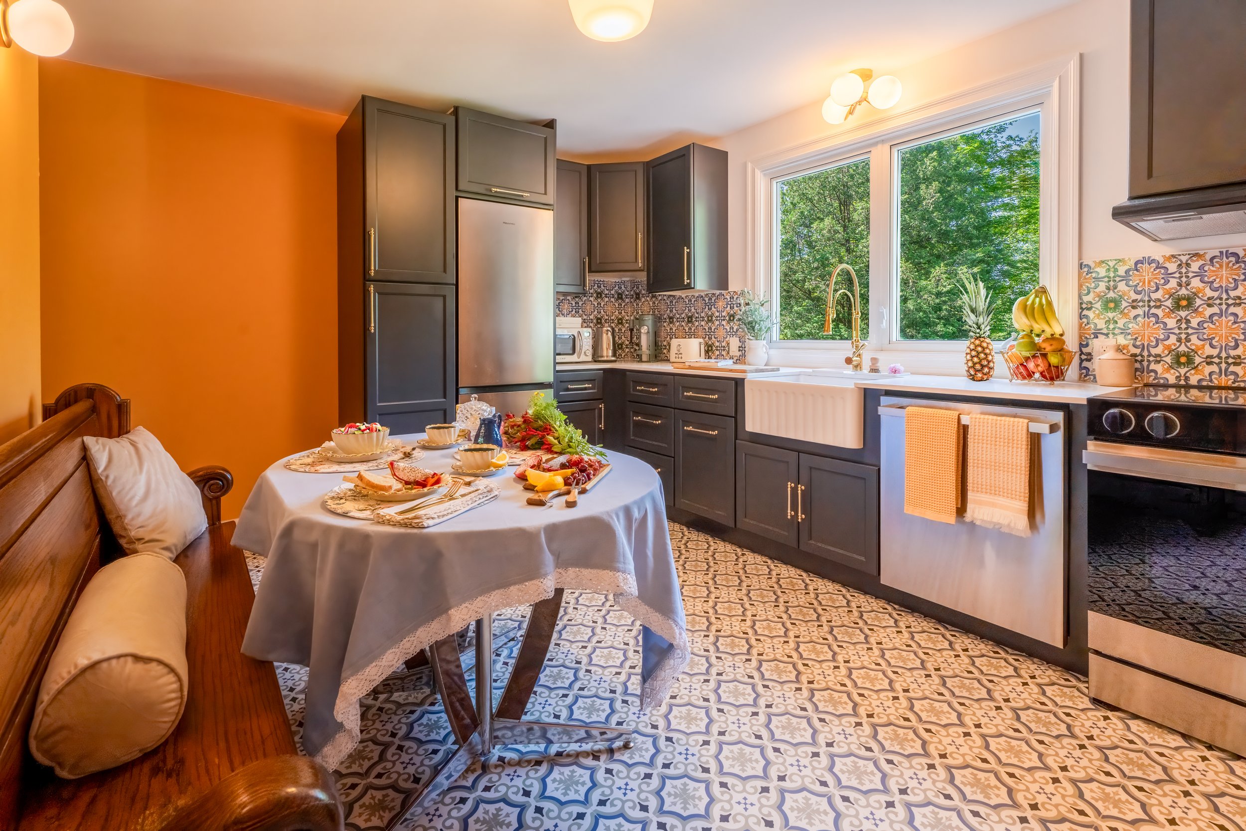 Kitchen with dark cabinets, patterned backsplash, large window with greenery outside, sink, fruit bowl, and a dining table with a tablecloth and breakfast items.