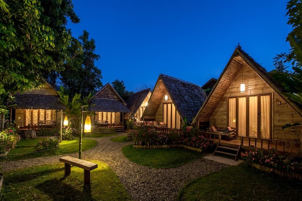 Night view of rustic wooden cottages with steep roofs, lit by warm outdoor lighting, surrounded by trees, plants, and a gravel pathway.