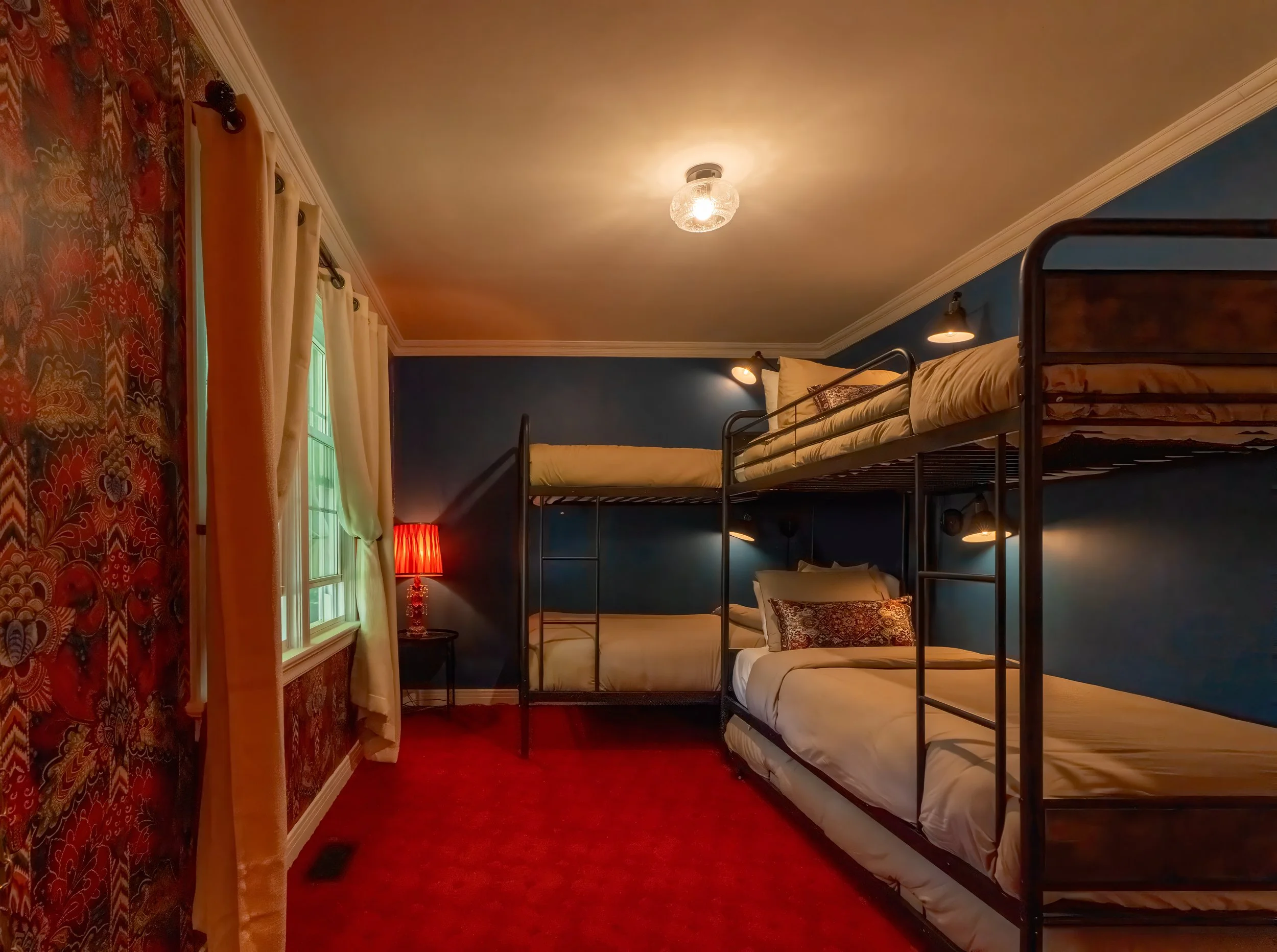 A bedroom with blue walls, a window with white curtains, a red lamp on a small table, and a black metal bunk bed with white bedding and decorative pillows, red carpet, and ceiling lights.
