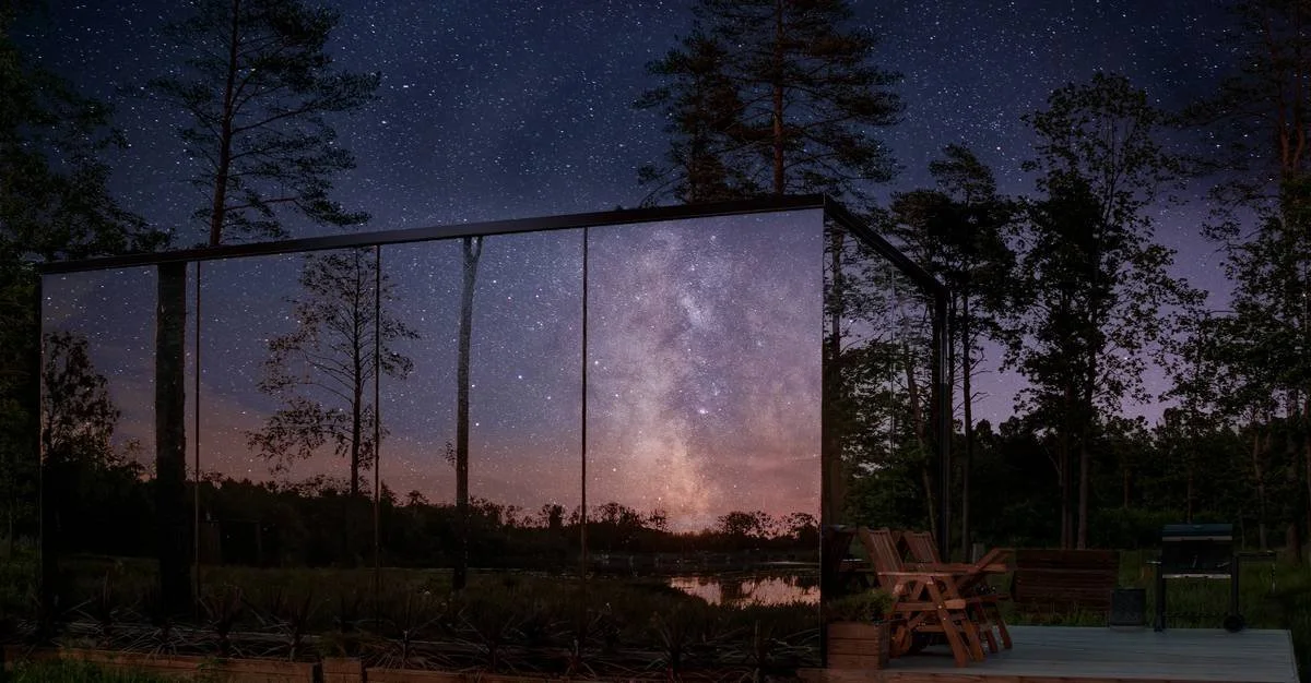 Nighttime outdoor scene featuring a large glass mirror reflecting the starry sky and Milky Way, with trees and a wooden deck with chairs and a grill.