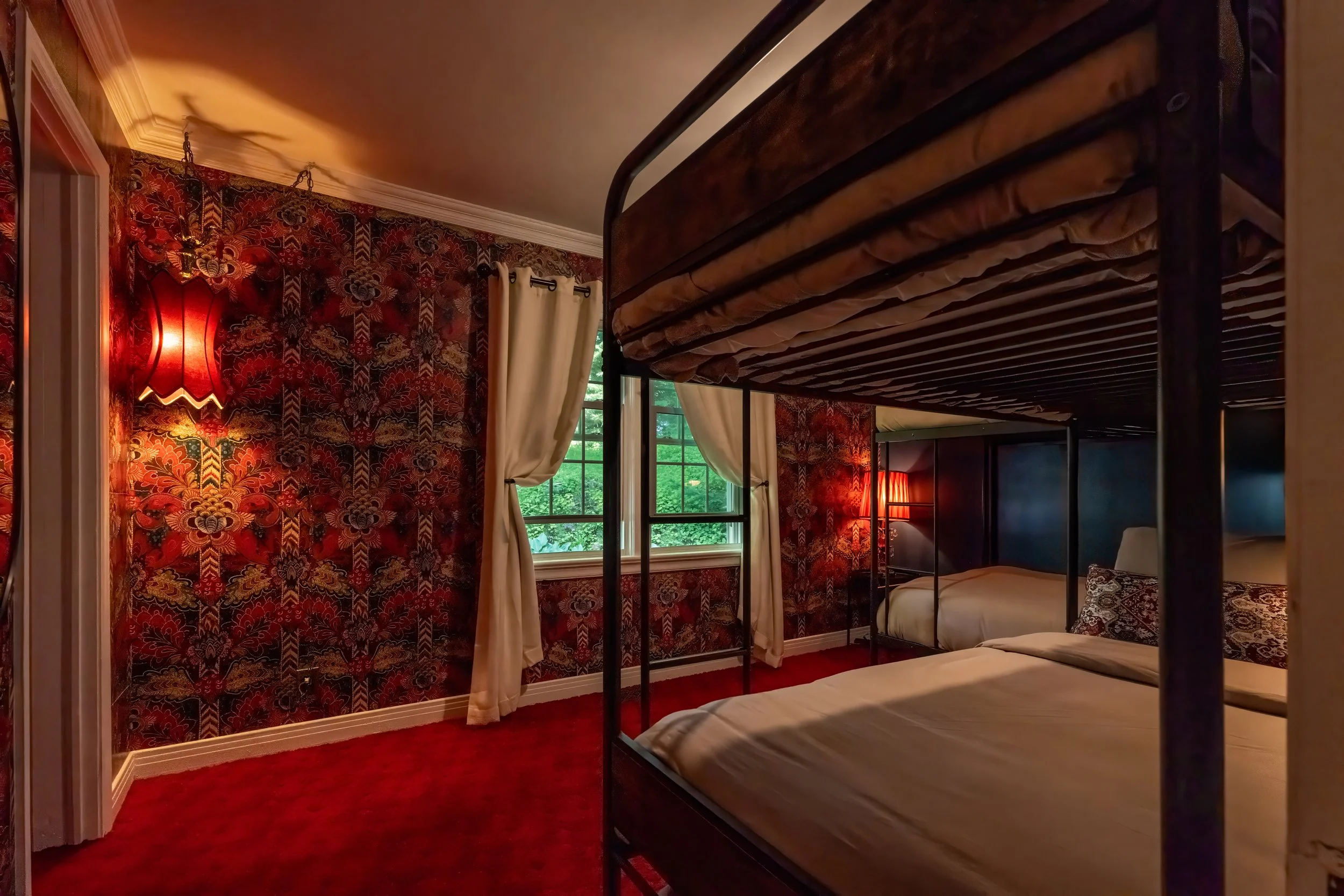 A bedroom with red patterned wallpaper, cream curtains, and two windows showing green trees outside. There is a black metal bunk bed with beige bedding and pillows, and two red lamps providing warm lighting.