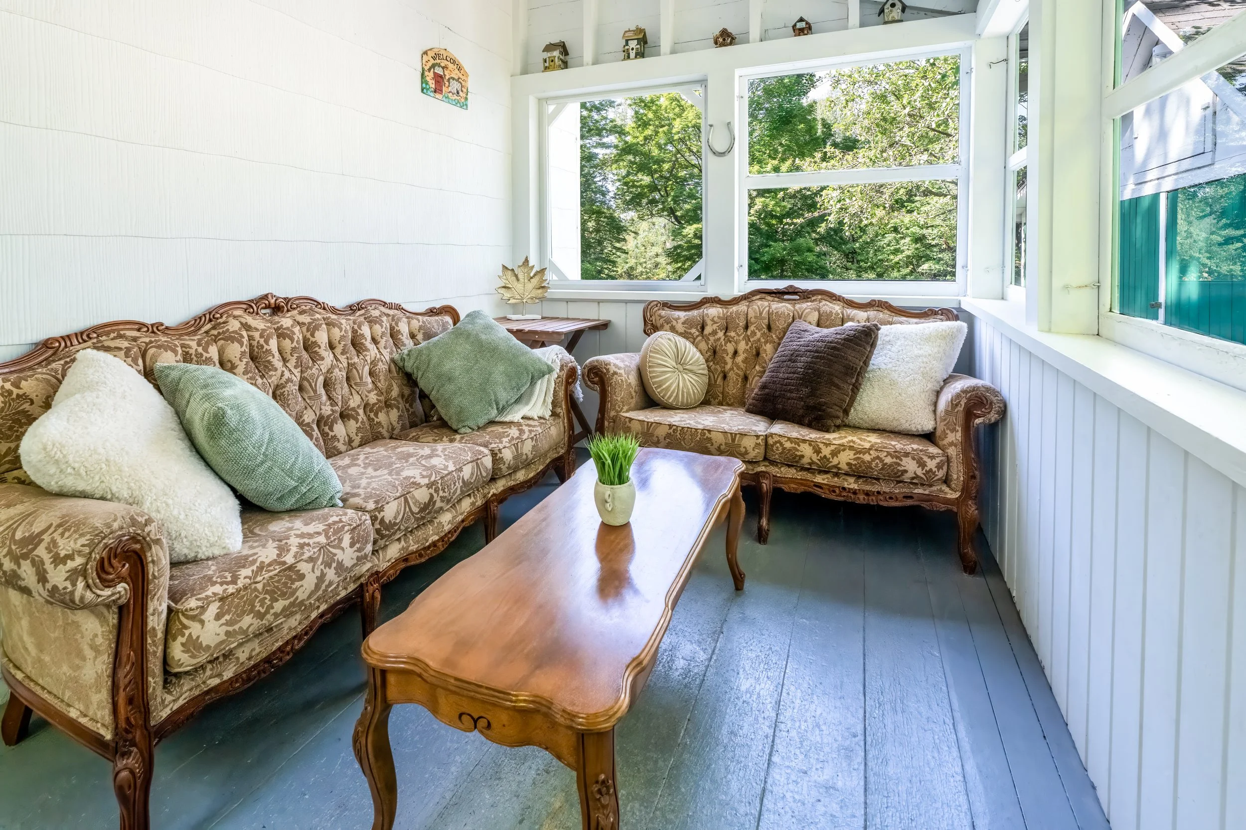 A cozy sunroom with two vintage floral sofas, multiple throw pillows, a wooden coffee table with a small plant, large windows letting in natural light, outdoor greenery visible outside, and simple wall decor.