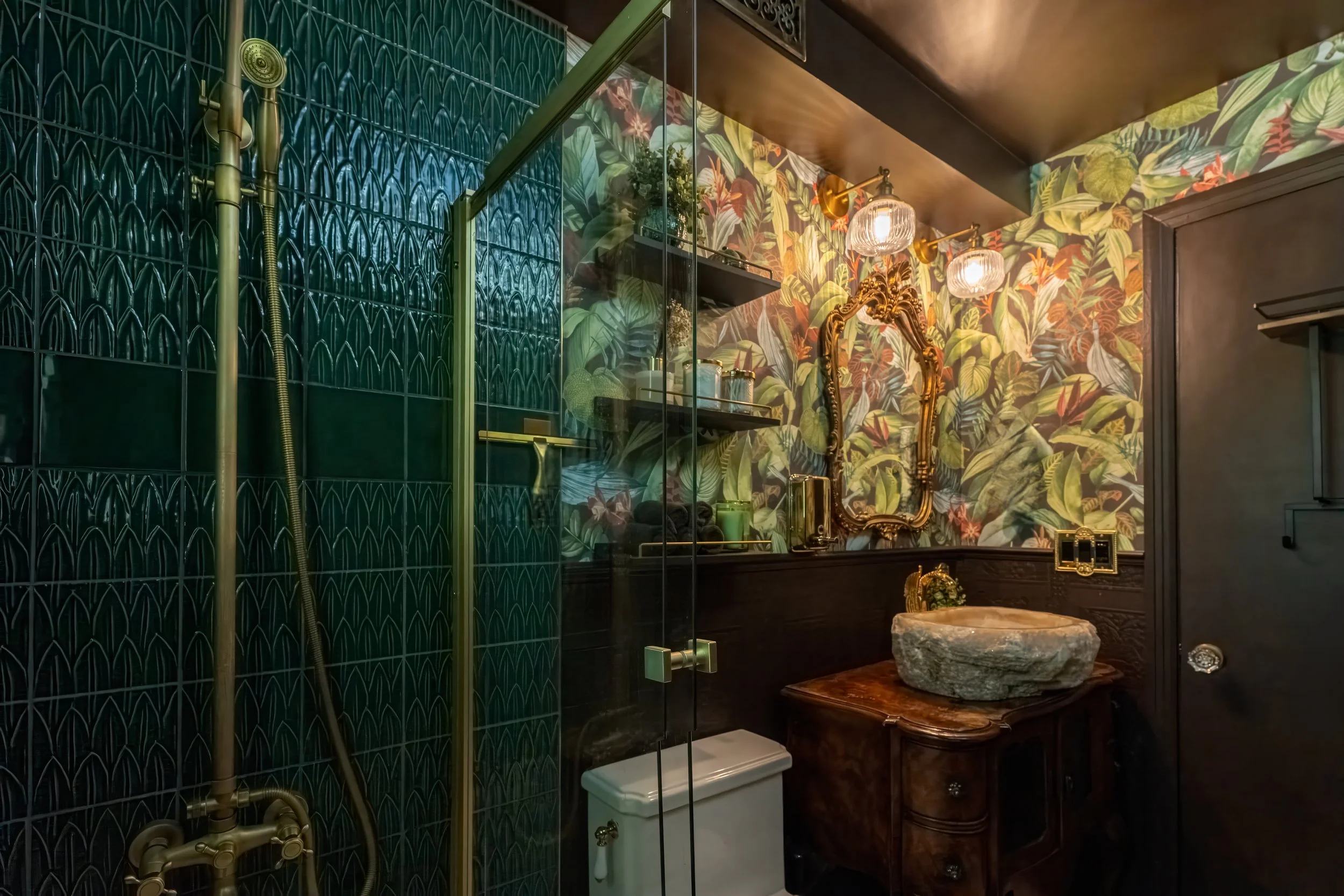 Bathroom with dark green tiled shower area, floral wallpaper, vintage mirror, stone sink, wooden vanity, and decorative lighting.