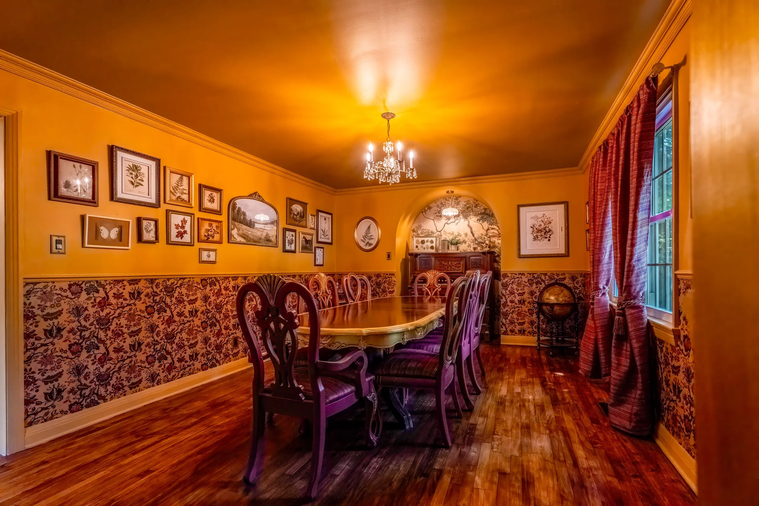 A dining room with yellow walls, a vintage chandelier, a large wooden dining table with chairs, framed botanical artwork, a window with red curtains, and antique decor.